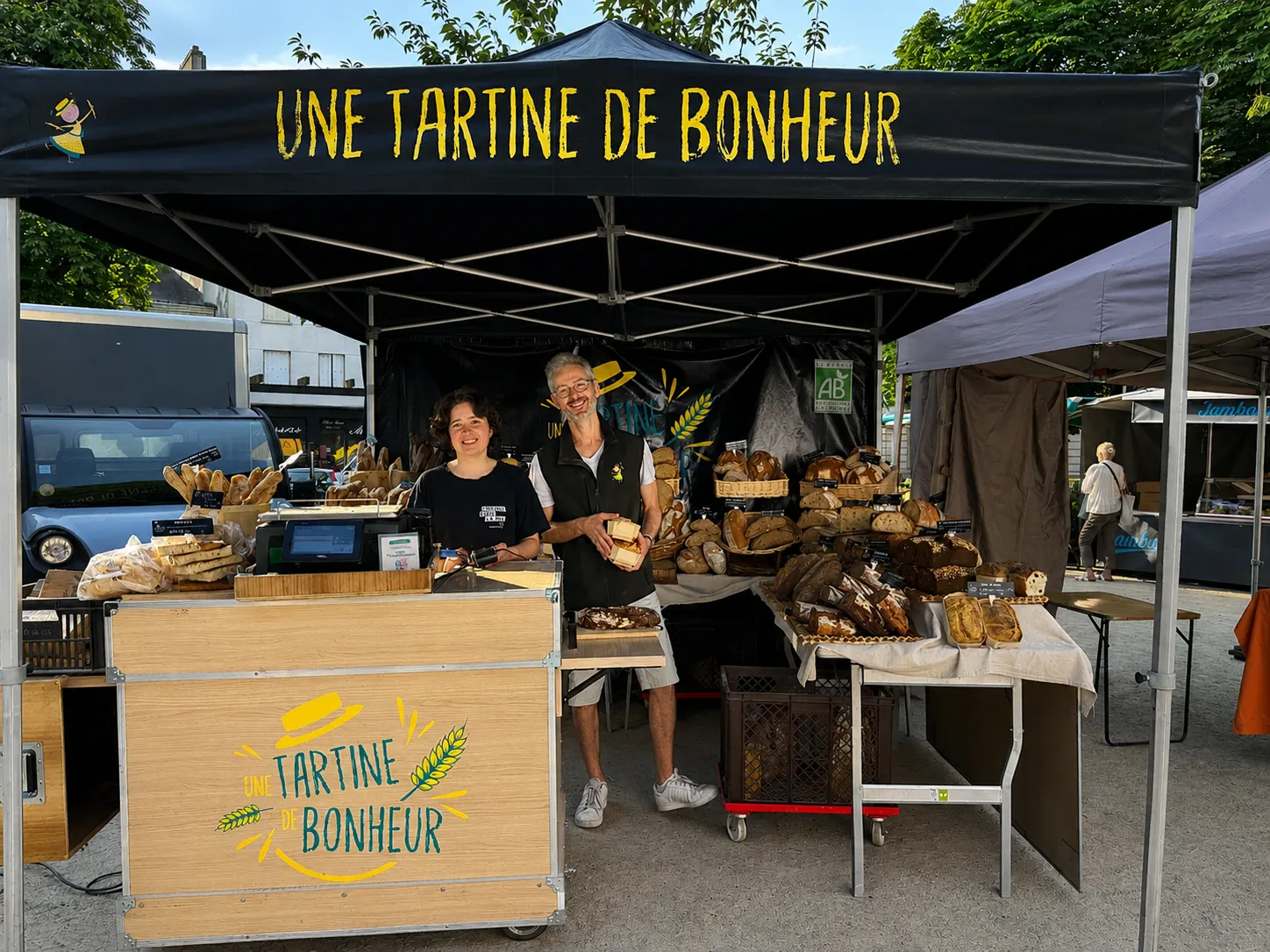 Deux vendeurs souriants dans un stand de marché avec divers pains sous une tente noire.