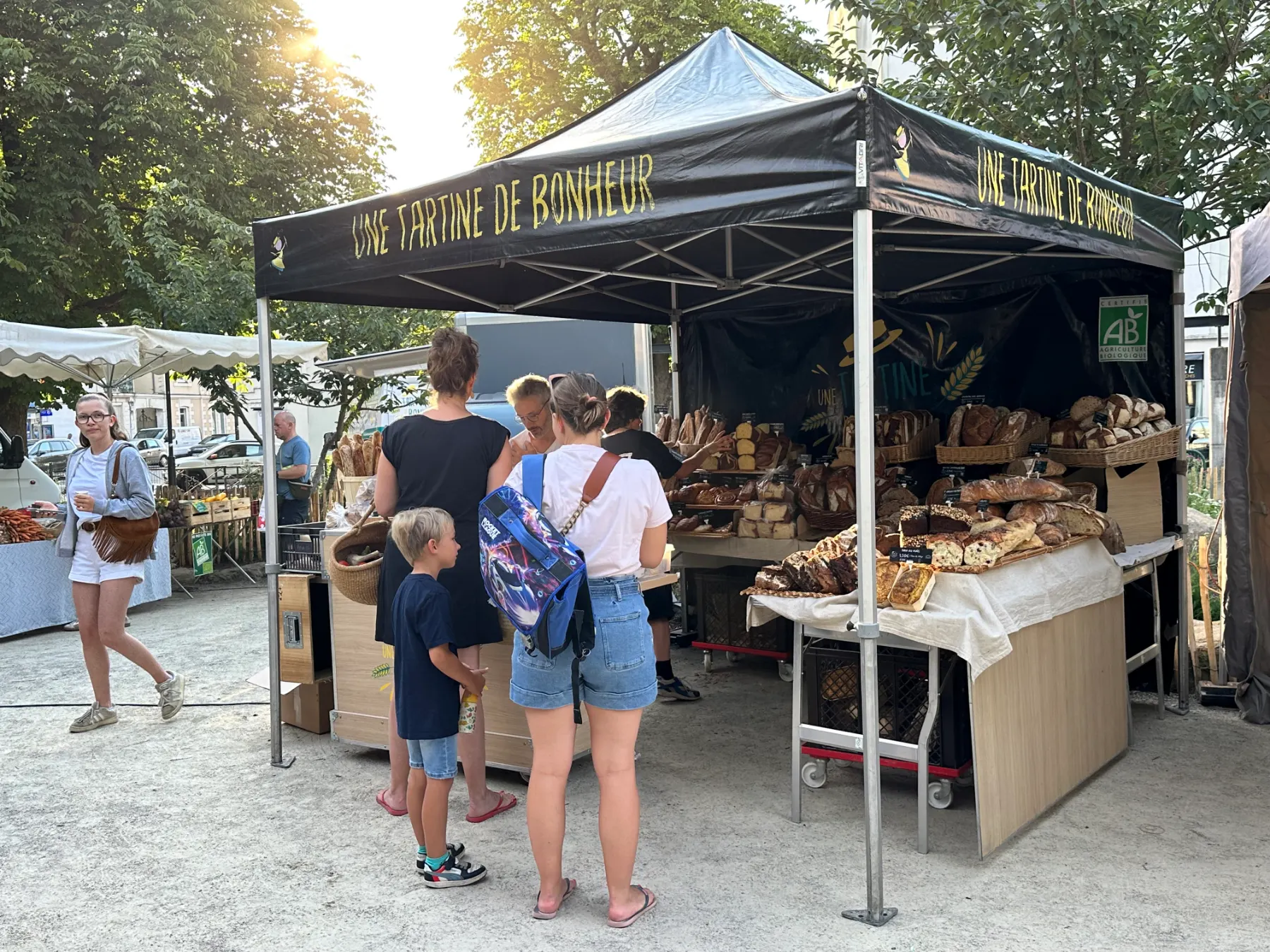 Marché en plein air avec stand de pain bio intitulé Une Tartine de Bonheur et clients faisant leurs achats.