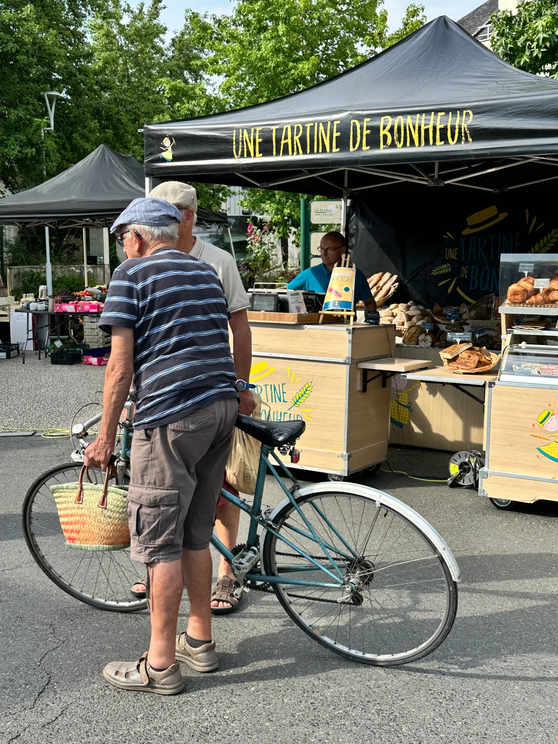 Deux hommes discutent devant un stand de pain intitulé Une Tartine de Bonheur, l’un avec un vélo.