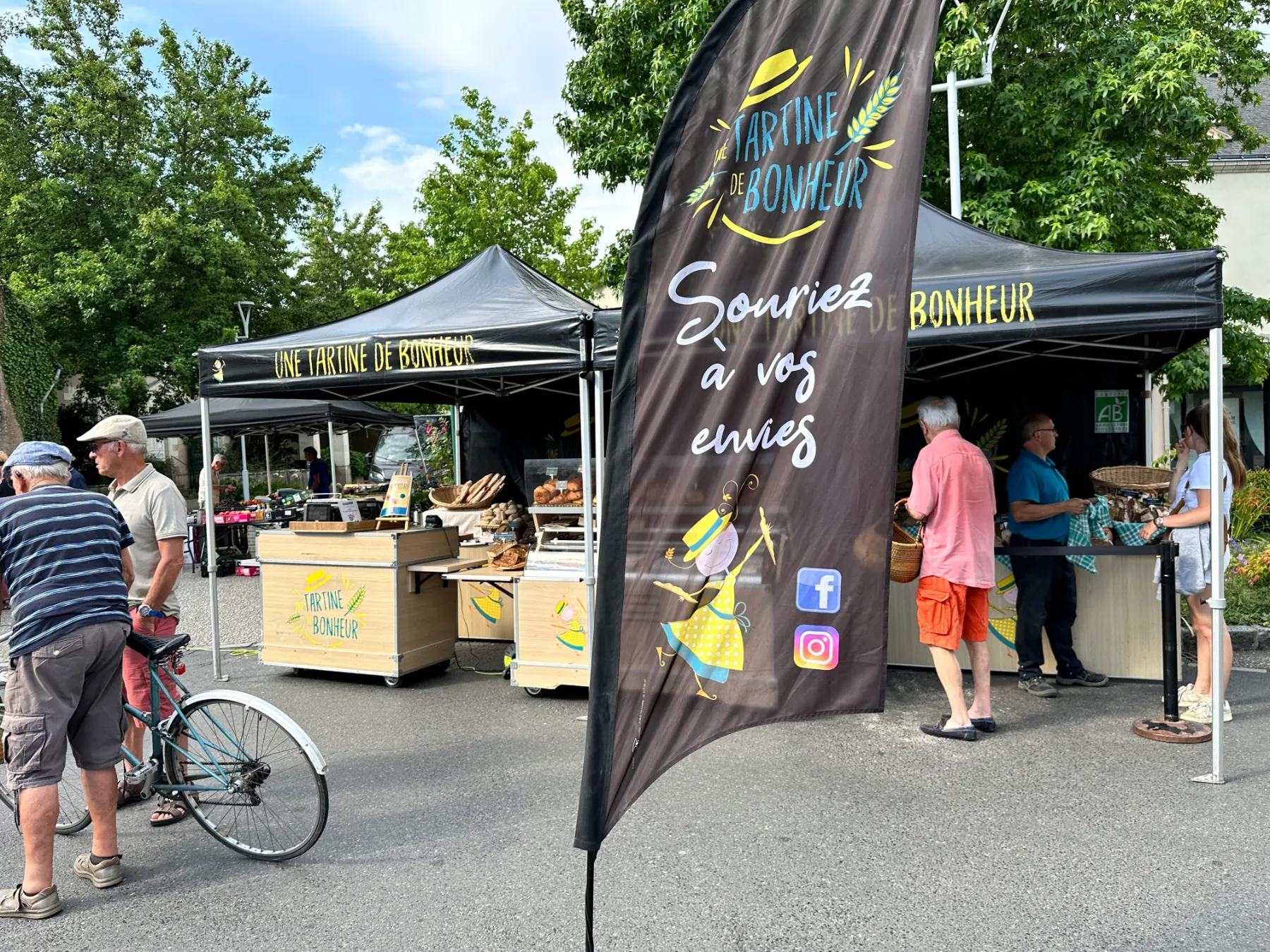 Stand de marché en plein air nommé Une Tartine de Bonheur avec des clients et une bannière noire.