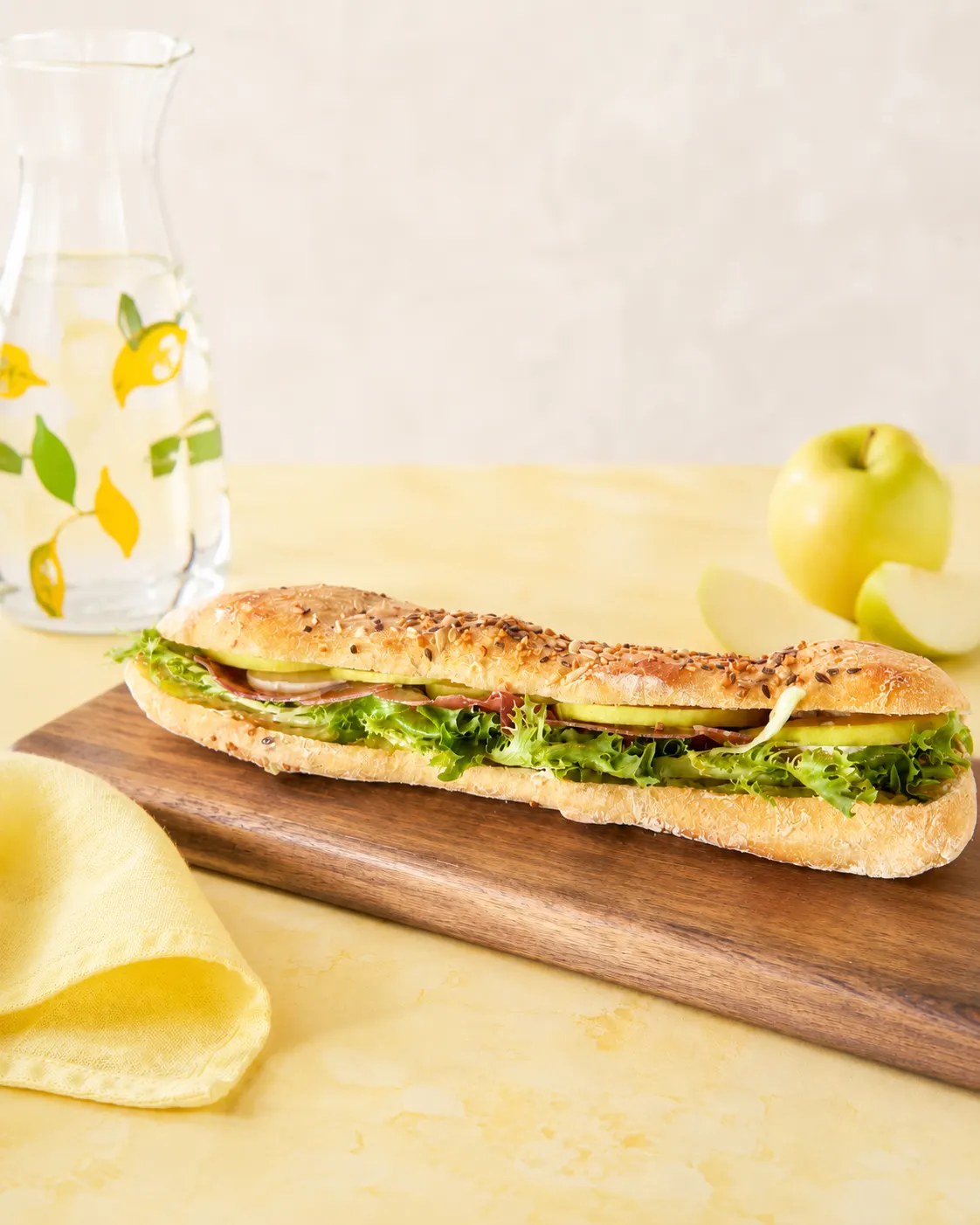 Sandwich ciabatta aux graines avec laitue, tranches de pomme verte et jambon sur une planche en bois.