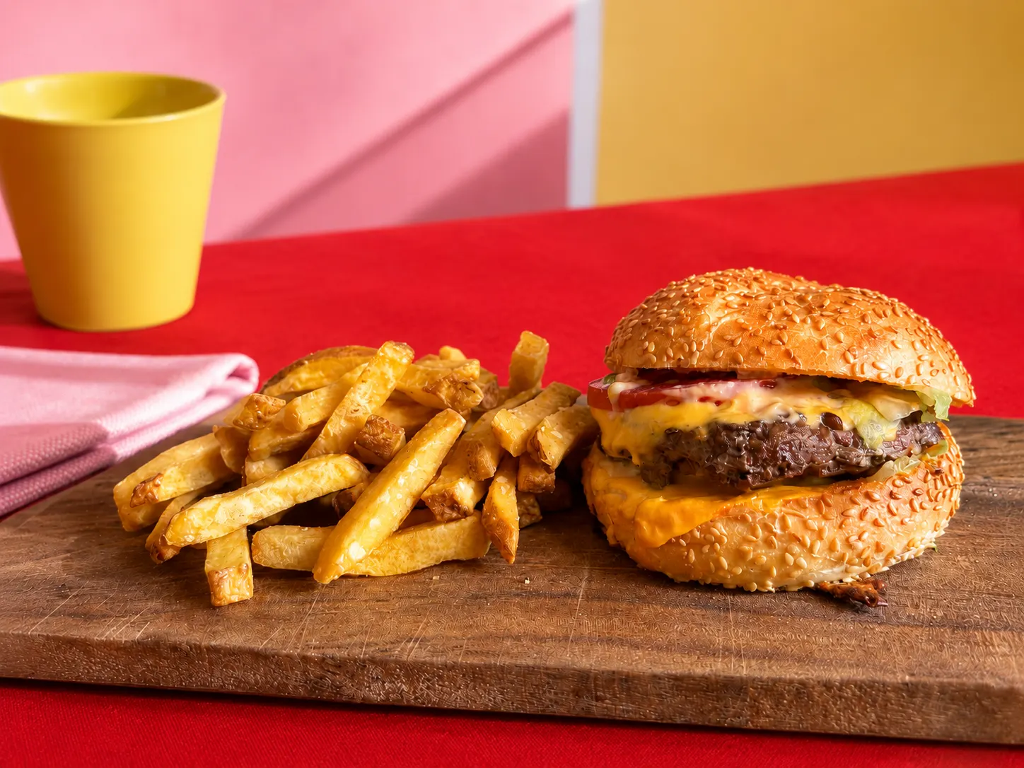 Un cheeseburger avec frites sur une planche en bois, fond rouge avec une tasse jaune.