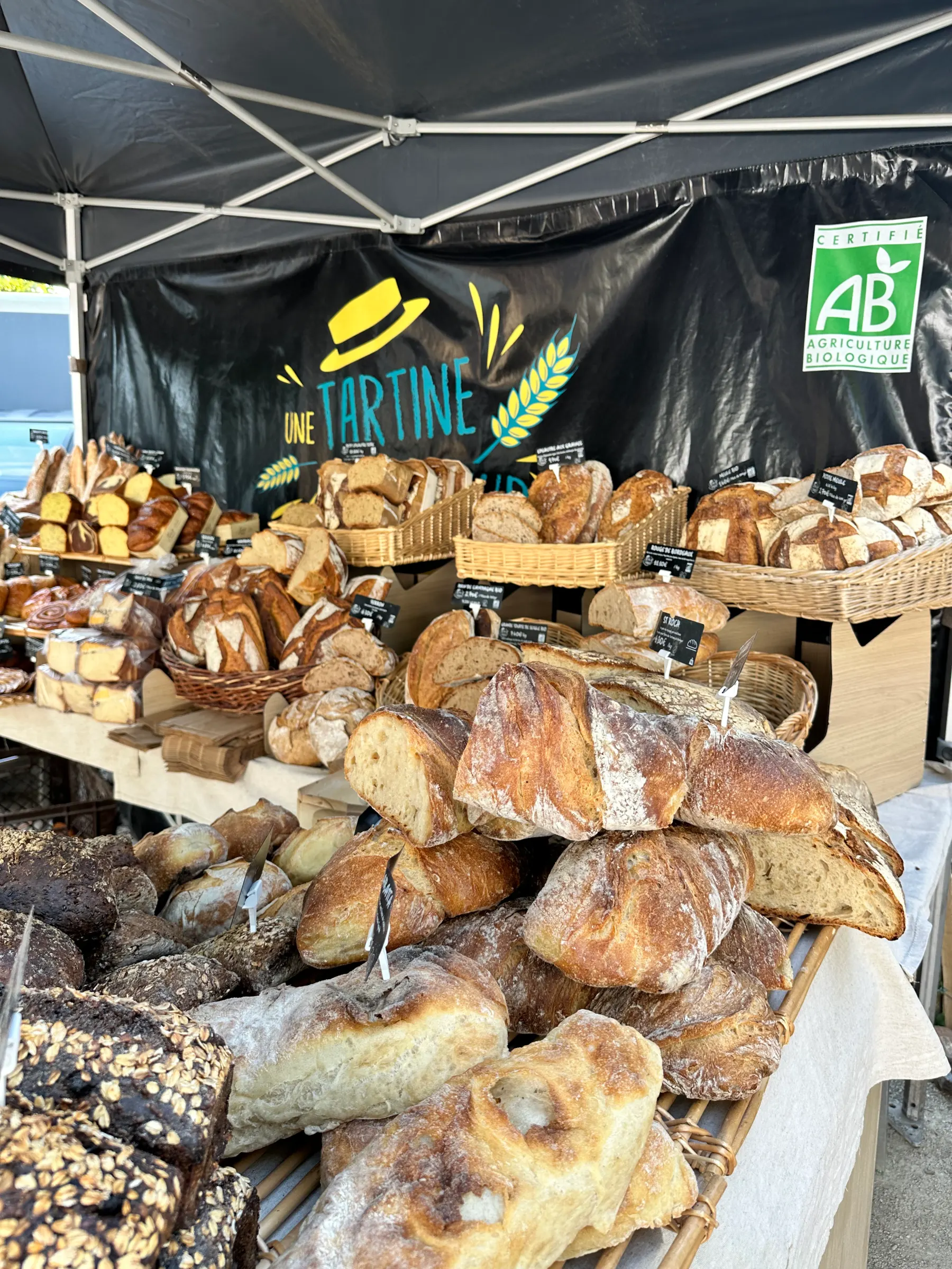 Étals de marché avec plusieurs pains variés sous tente, bannière agriculture biologique visible.