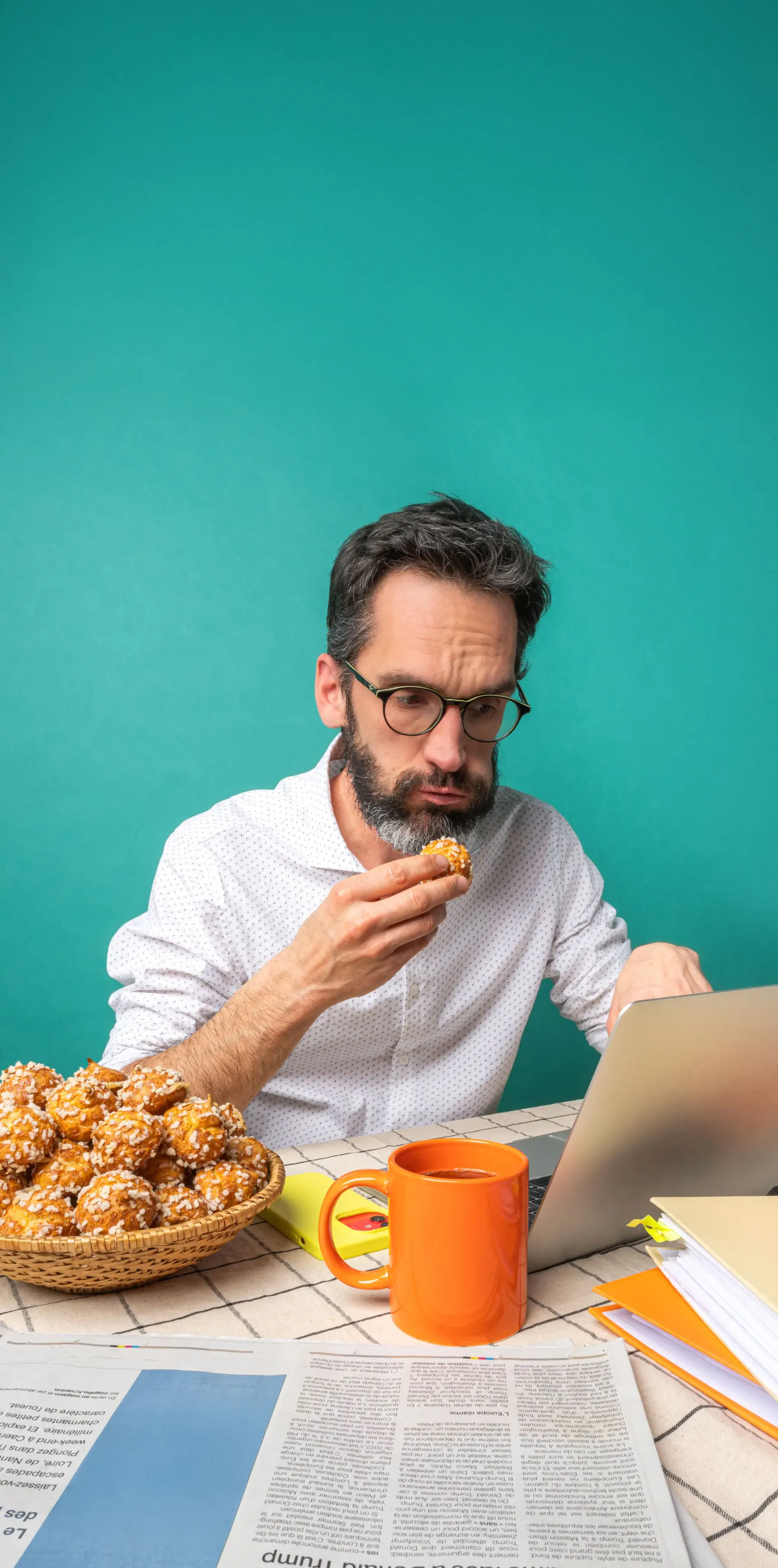 Homme portant des lunettes mangeant un beignet près d'un ordinateur portable et une tasse orange.