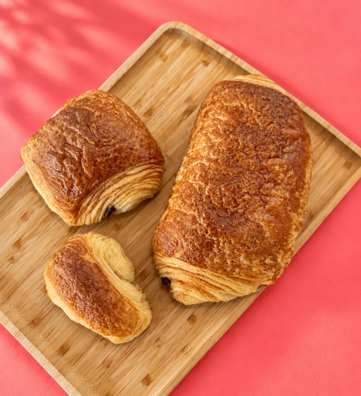Trois viennoiseries feuilletées dorées sur un plateau en bois posé sur une surface rose vif.