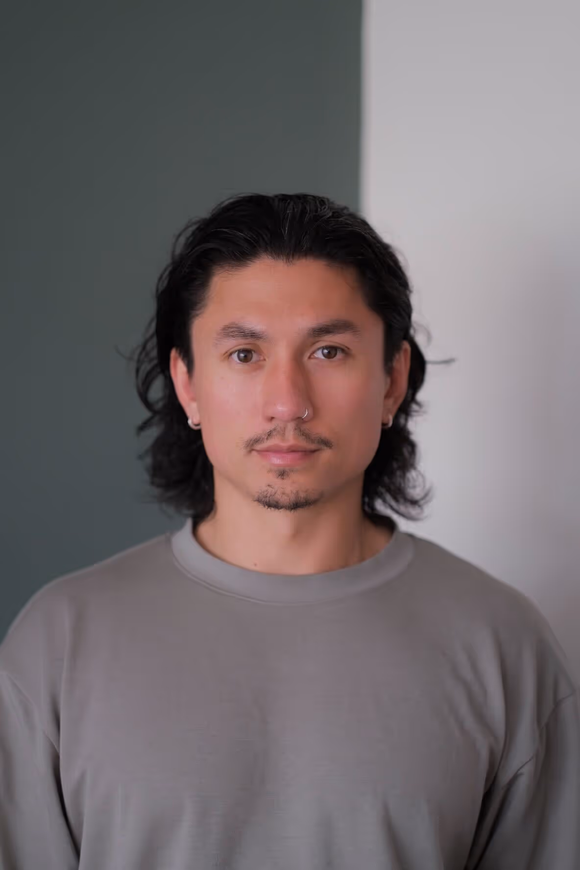 Portrait of a young man with medium-length dark hair, light facial hair, nose ring, and earrings, wearing a gray shirt, standing in front of a wall divided into dark green and light gray sections.