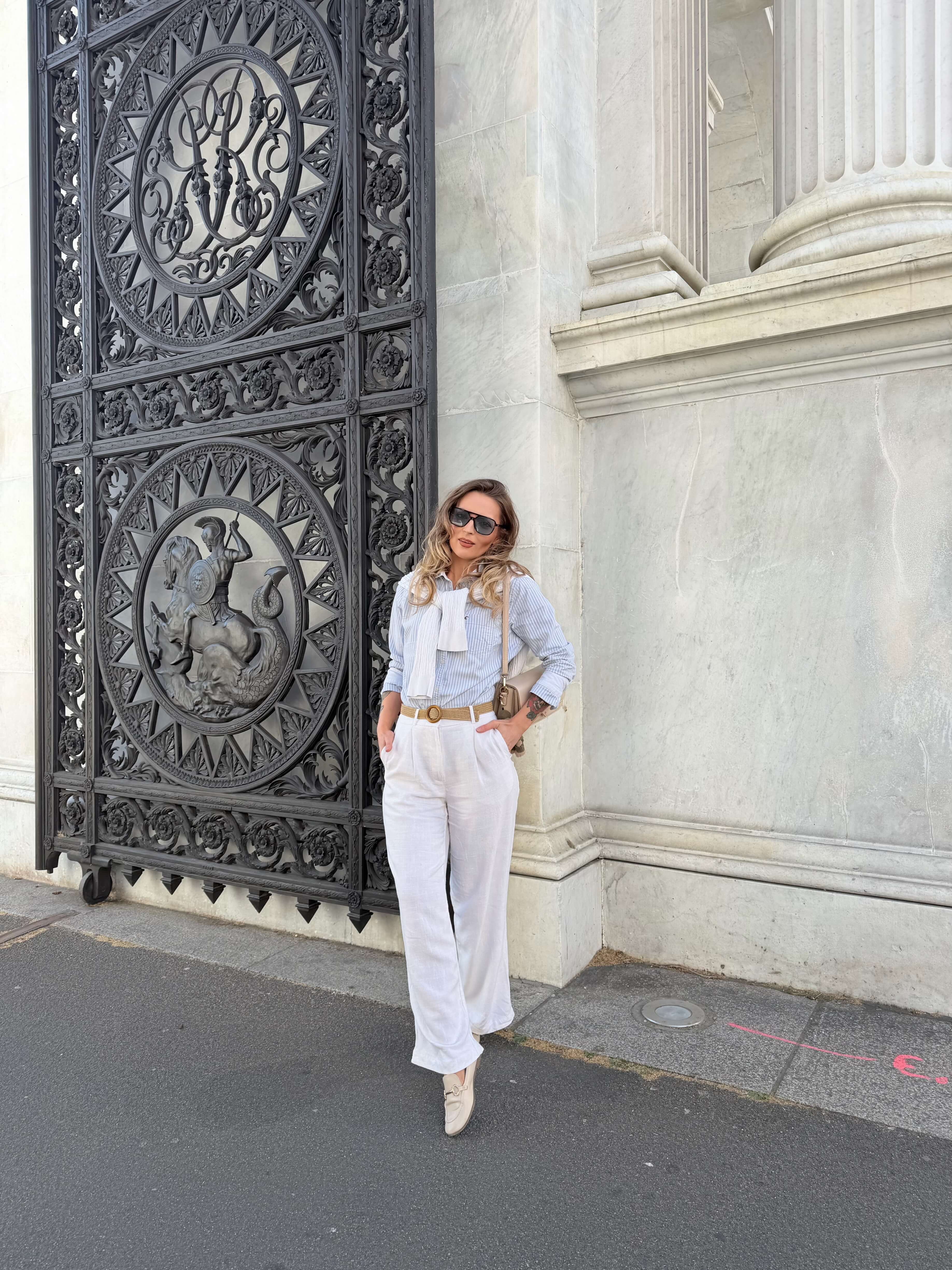Woman wearing sunglasses, light blue striped shirt with white sweater draped over shoulders, white pants, and beige shoes leaning against a marble building near an ornate black metal gate with sculpted circular designs.