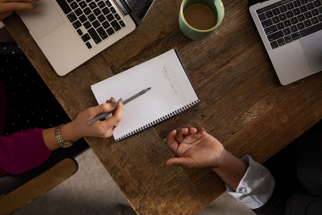 A woman writes on a notepad while two men work on laptops at a table, engaged in a collaborative discussion
