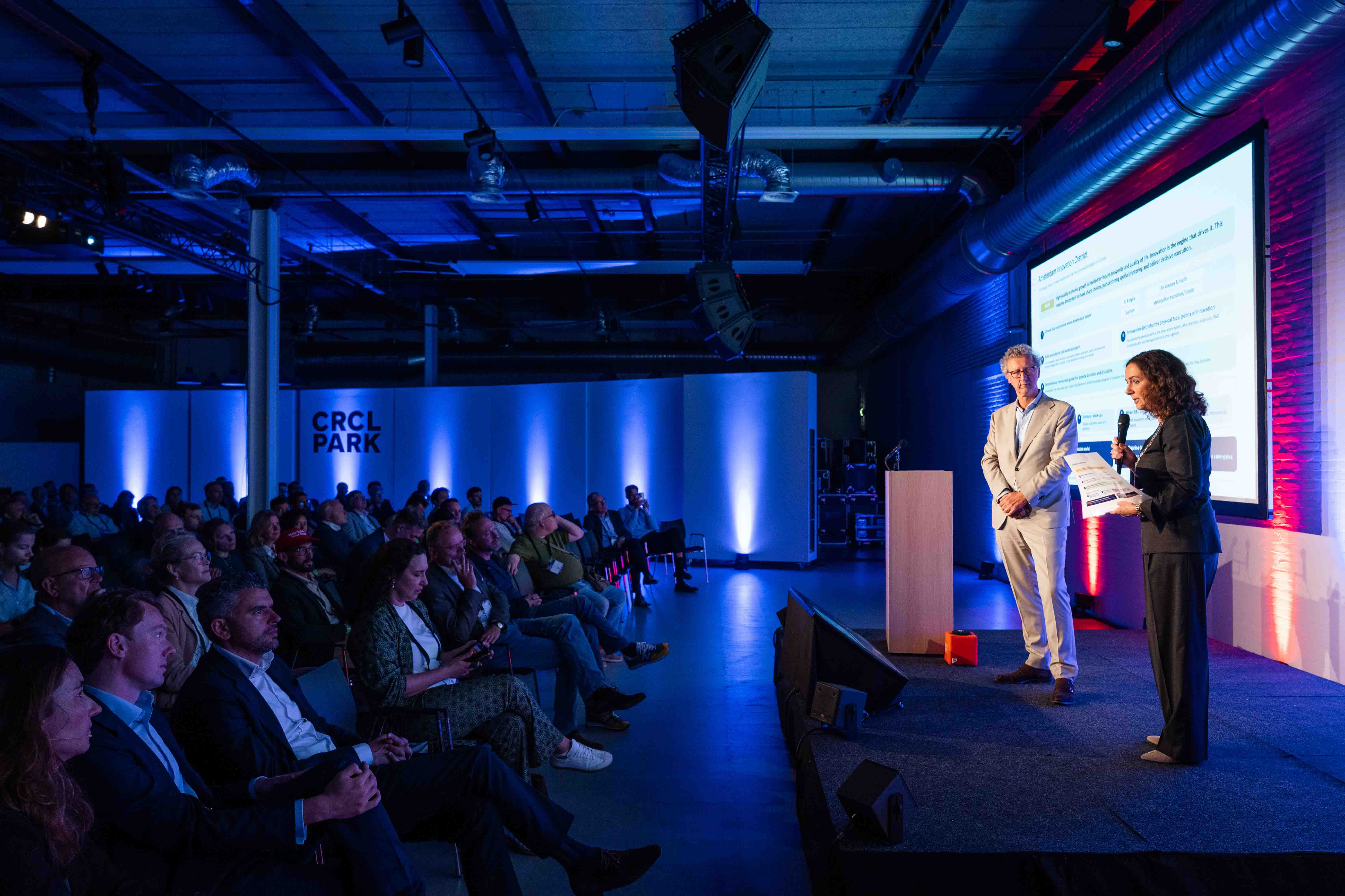 Interview op het podium met scherm op de achtergrond tijdens de Amsterdam Deep Tech Showcase - Sennheiser EWDX handheld