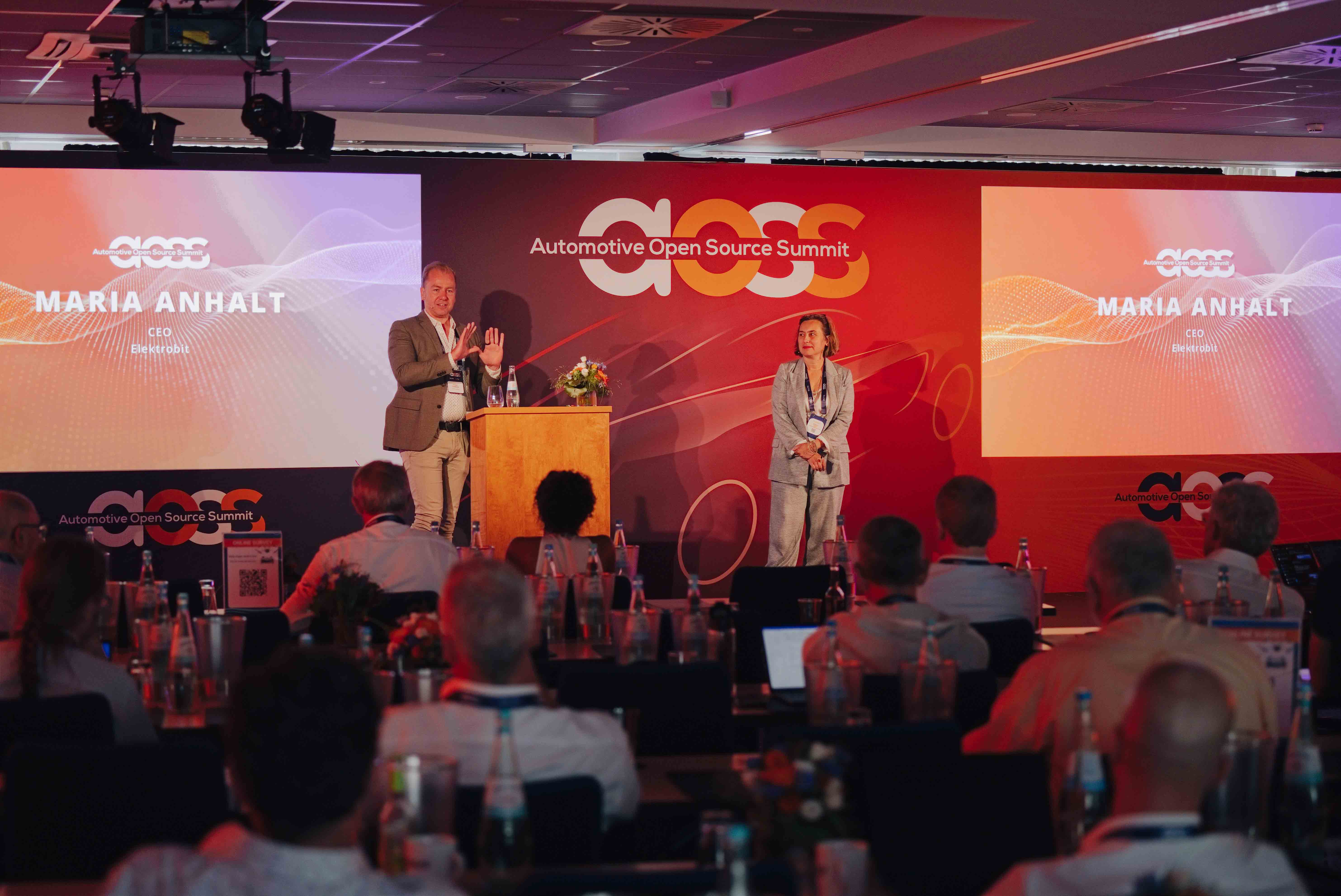 Twee sprekers introduceren een sessie op het podium van de Automotive Open Source Summit in Starnberg met projectieschermen op de achterwand