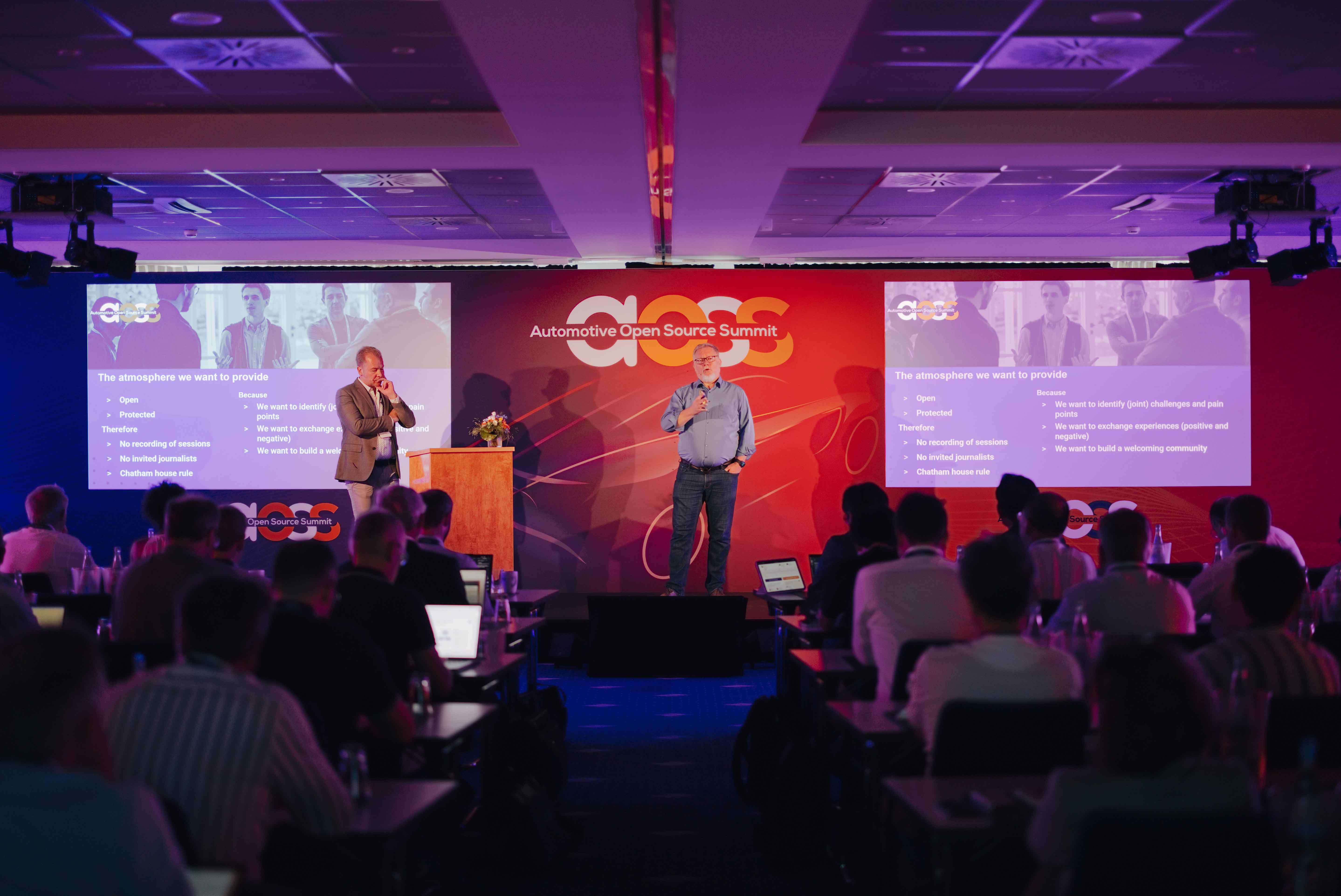 Spreker op het podium met de brede projectiewand tijdens de Automotive Open Source Summit in Starnberg