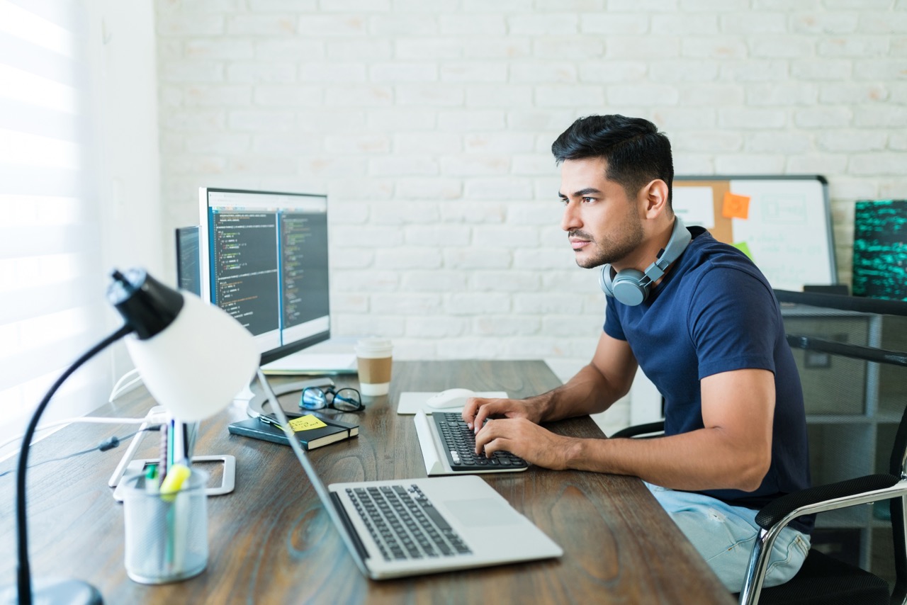 A male student at a desk typing on a laptop with a monitor displaying code.