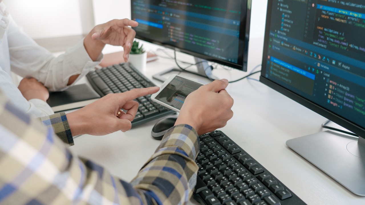 Two people reviewing code on a smartphone and dual monitors at a desk.