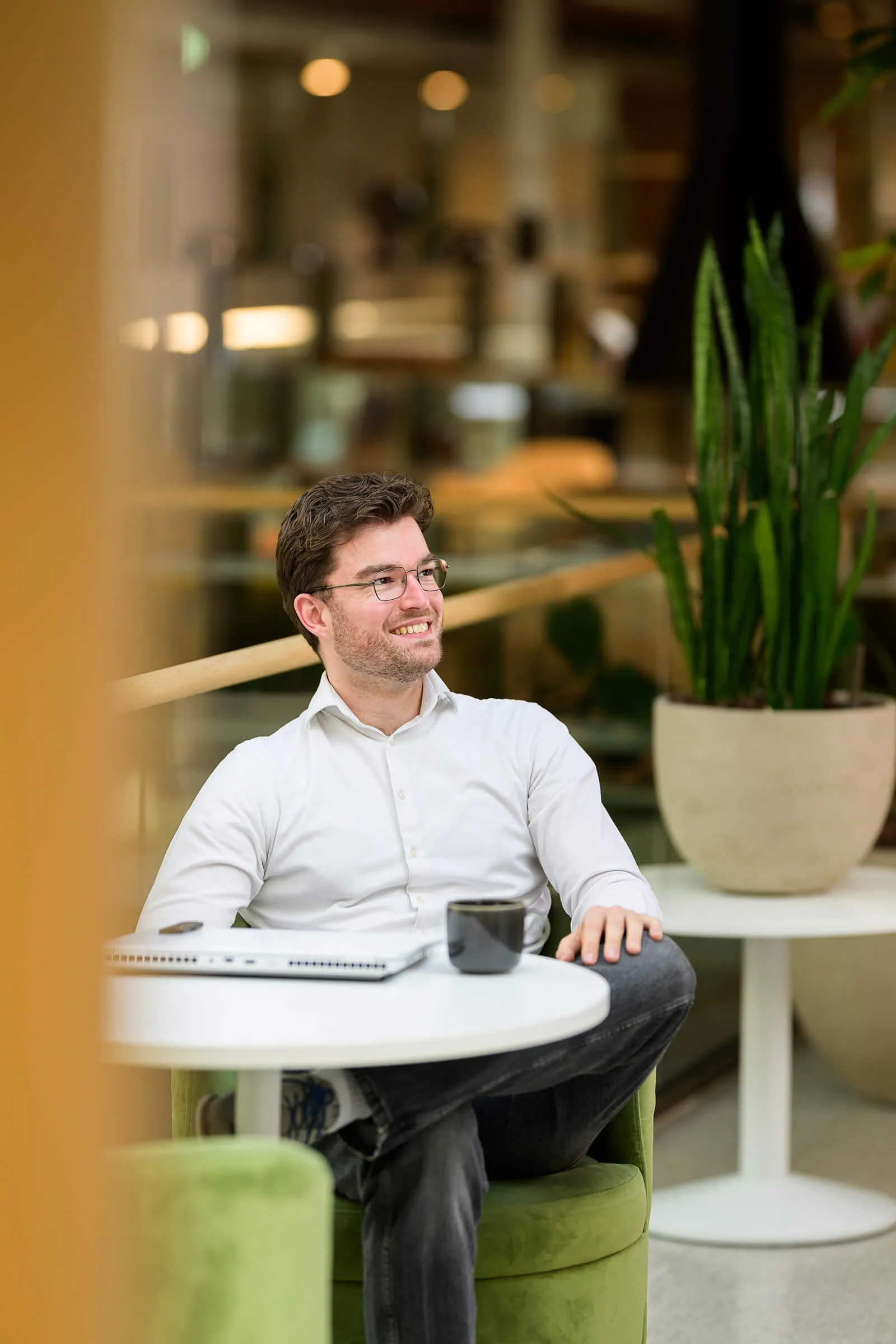 Arxion-consultant met bril zit lachend aan een tafeltje met laptop en koffie