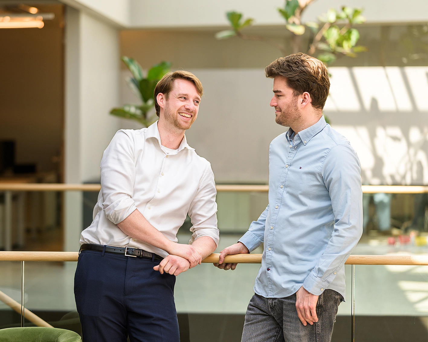 Twee jonge mannen praten en lachen bij een balustrade in een lichte kantoorruimte.
