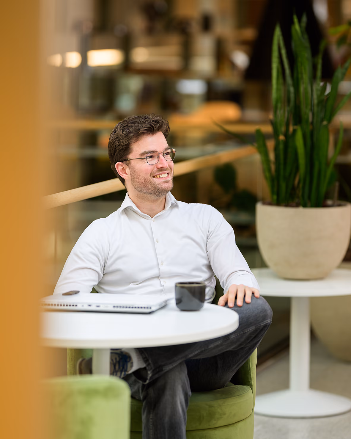 Arxion-consultant met bril zit lachend aan een tafeltje met laptop en koffie