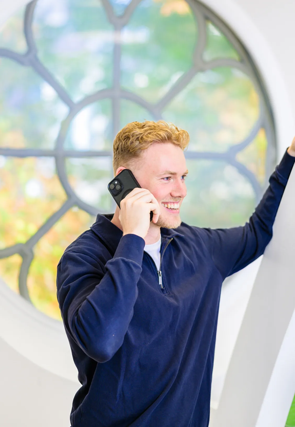 A young man with light brown hair wearing a navy blue sweater holds a smartphone to his ear while standing next to a large, decorative window.