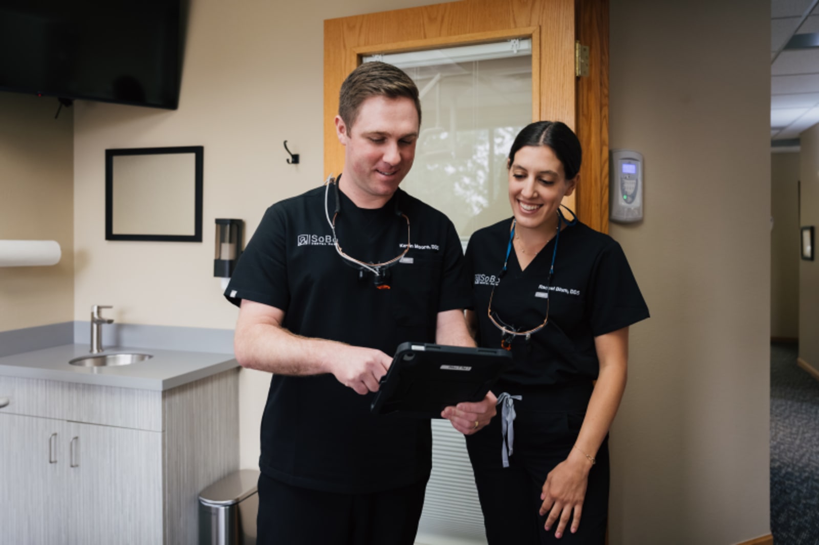 Two dental professionals in black scrubs smiling in a modern dental office treatment room.