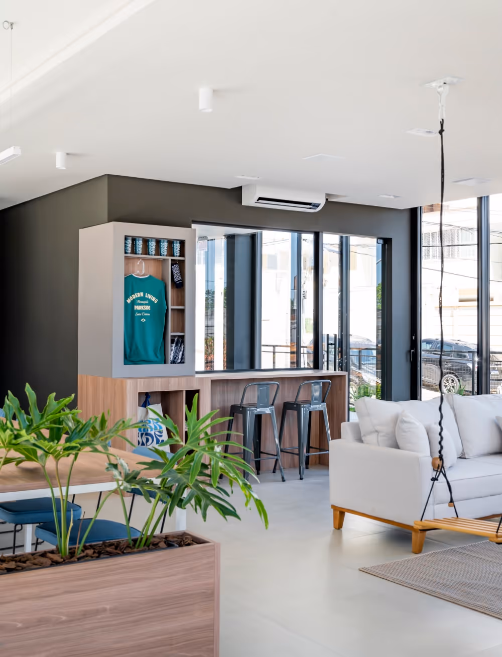 Sala moderna com sofá branco, plantas em vaso de madeira, banquetas metálicas e armário com camiseta verde pendurada.