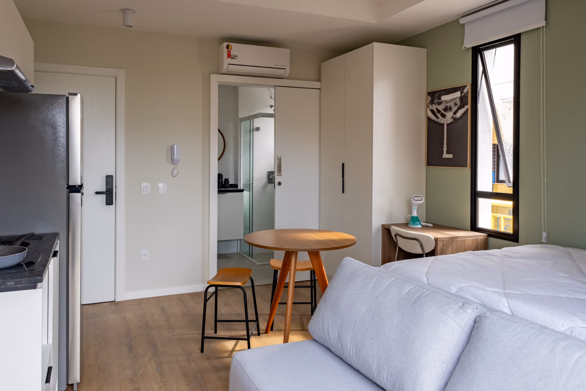 Sala de apartamento compacta com sofá cinza, mesa redonda de madeira com duas banquetas, dormitório com cama branca, guarda-roupa branco, escrivaninha de madeira ao lado da janela, cozinha com geladeira inox e bancada preta, e banheiro com porta de correr.