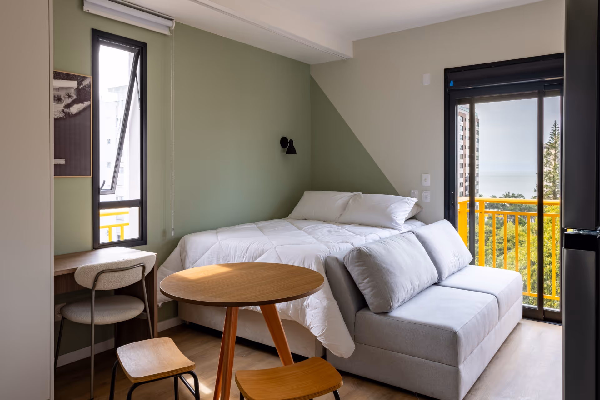 Quarto com cama branca, sofá cinza, mesa redonda de madeira com duas banquetas, escrivaninha com cadeira e sacada com vista para árvores e mar.