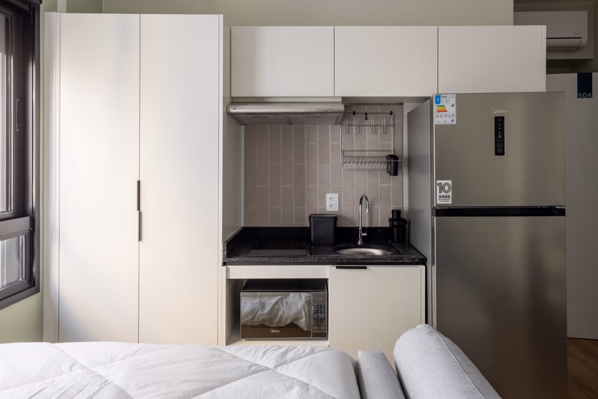Cozinha moderna com armários brancos, bancada preta, pia, micro-ondas, cooktop, exaustor e geladeira inox ao lado de uma janela e cama branca.