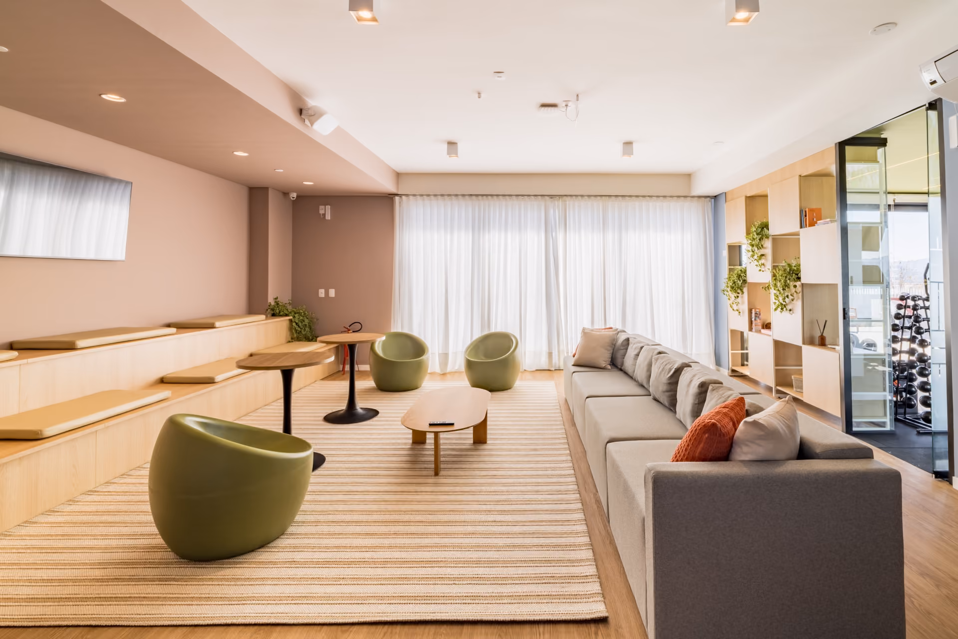 Sala de estar comprida com sofa cinza, poltronas verdes, arquibancada de madeira e estante ao fundo
