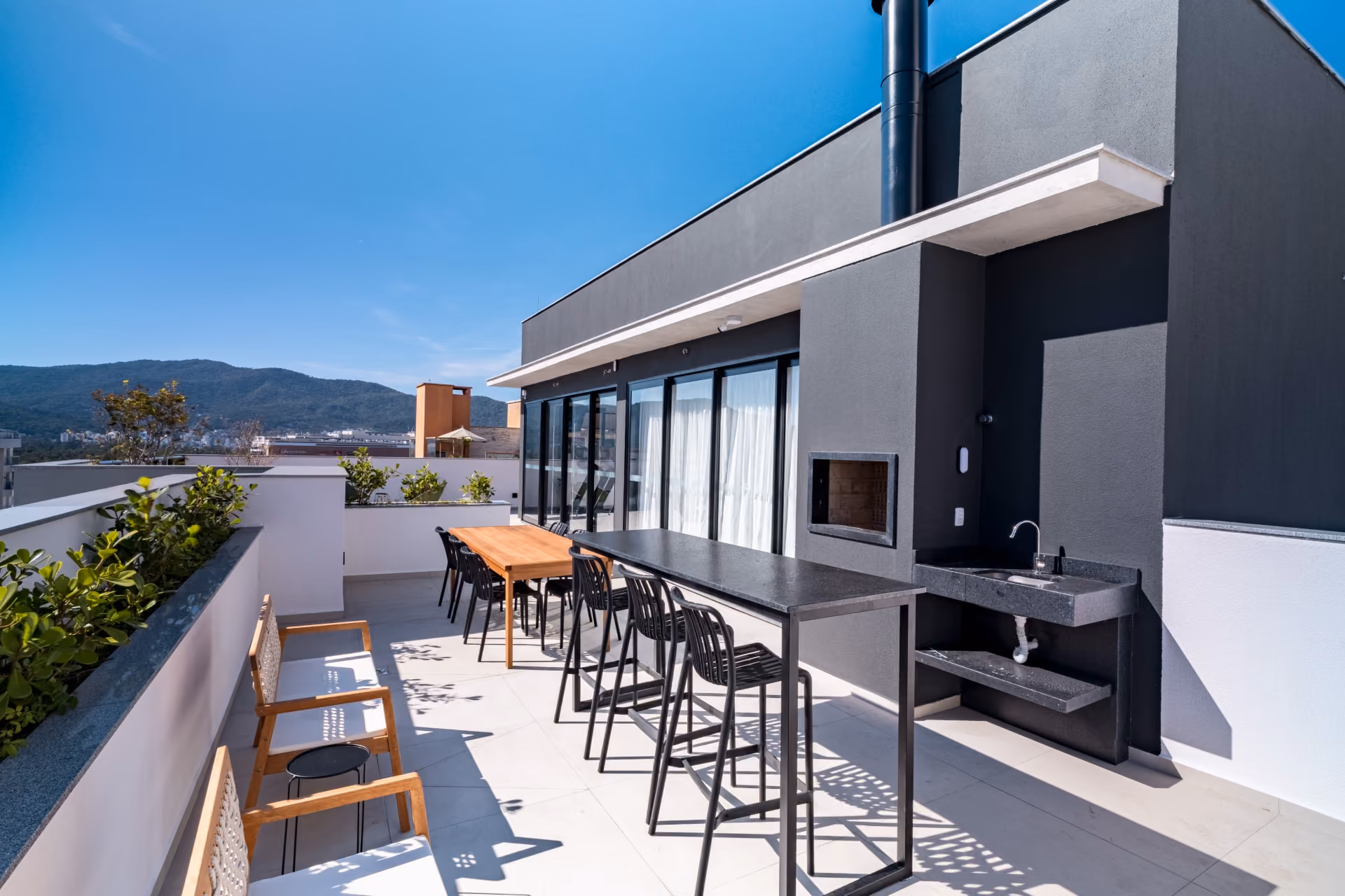 Terraco coletivo com churrasqueira, bancada preta comprida, banquetas e vista para os morros de Florianopolis
