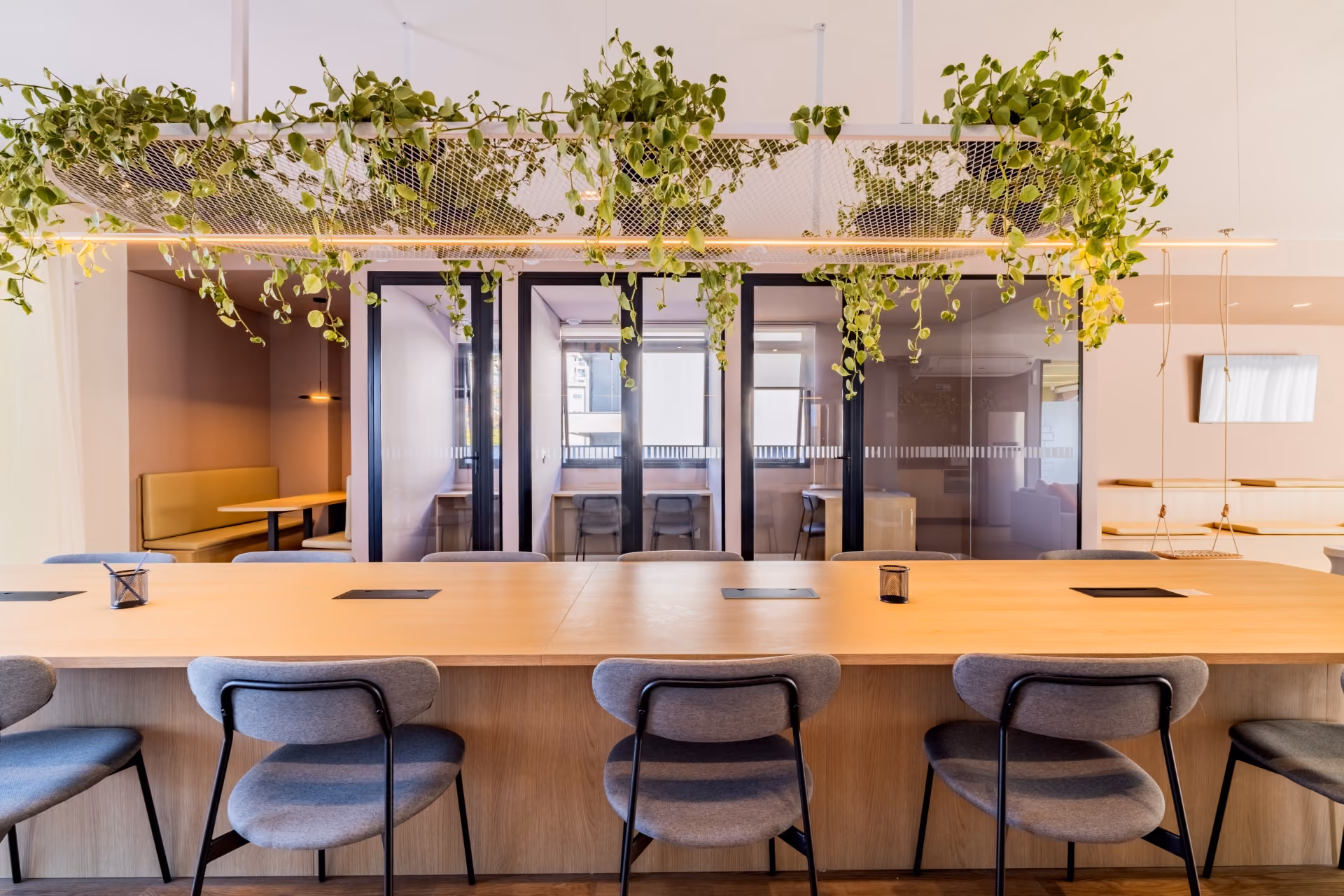 Mesa comunitaria do coworking sob jardim suspenso, com cadeiras estofadas e salas de reuniao ao fundo