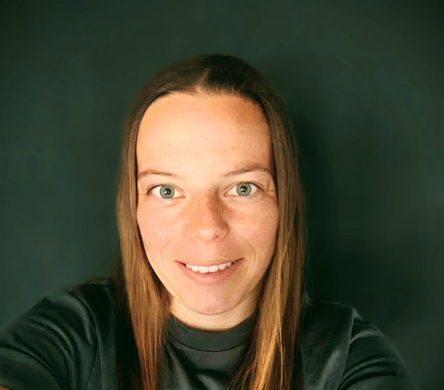 Marie von vorne bis zur Schulter vor einer dunklen Wand. Mit einem lächelnden Gesicht. Offene Haare. Neutrales, dunkles Shirt.