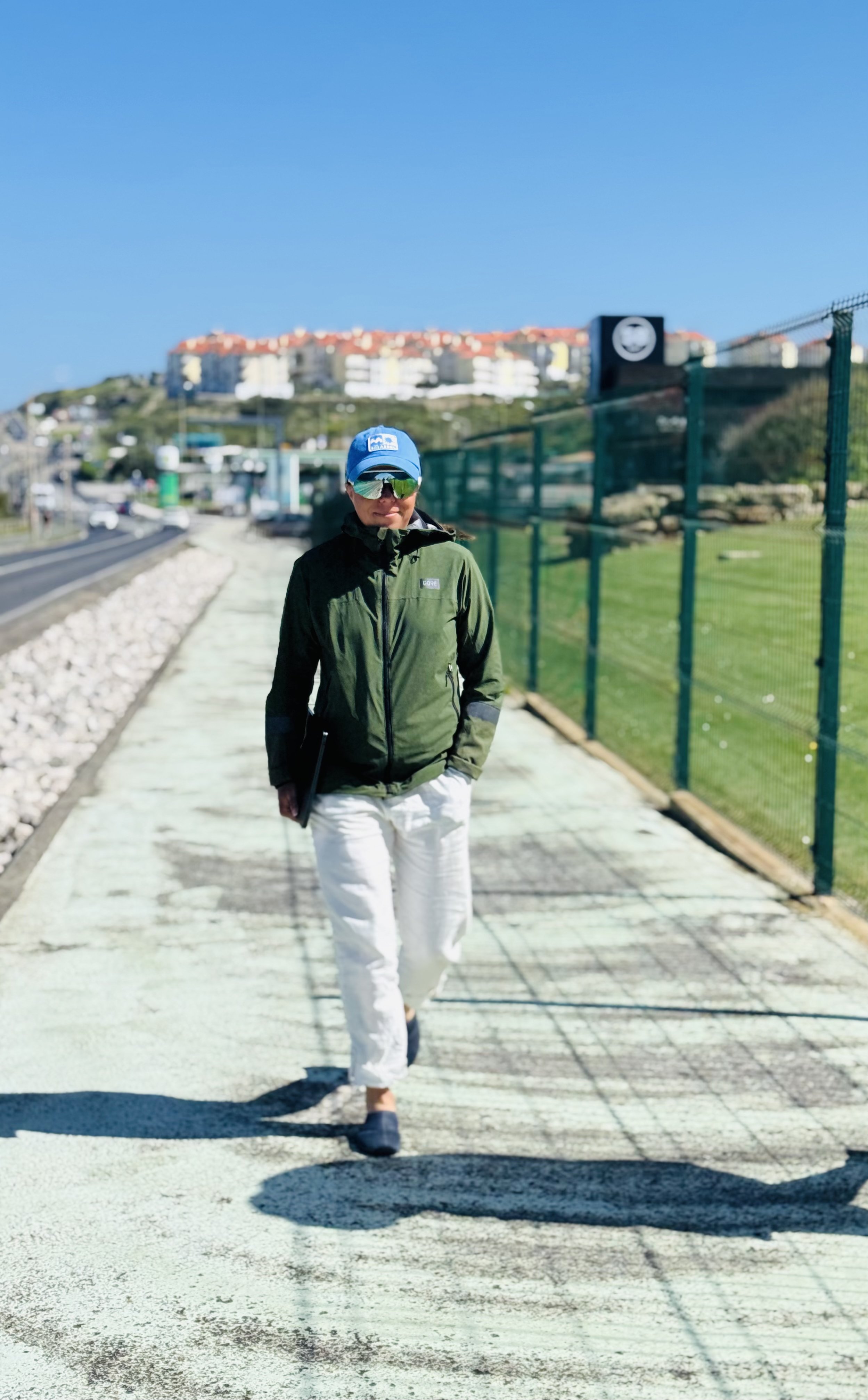 Marie von vorne in Portugal in Ericeira mit dem Laptop in der Hand auf einem Gehweg bei blauem Himmel. Mit weißer, langer Leinenhose, dunkelblauen Barfußschuhen, olive-grüner Windjacke von Gore, Fahrrad-Sonnebrille und hellblauem Billabong Surfcap auf dem kopf.