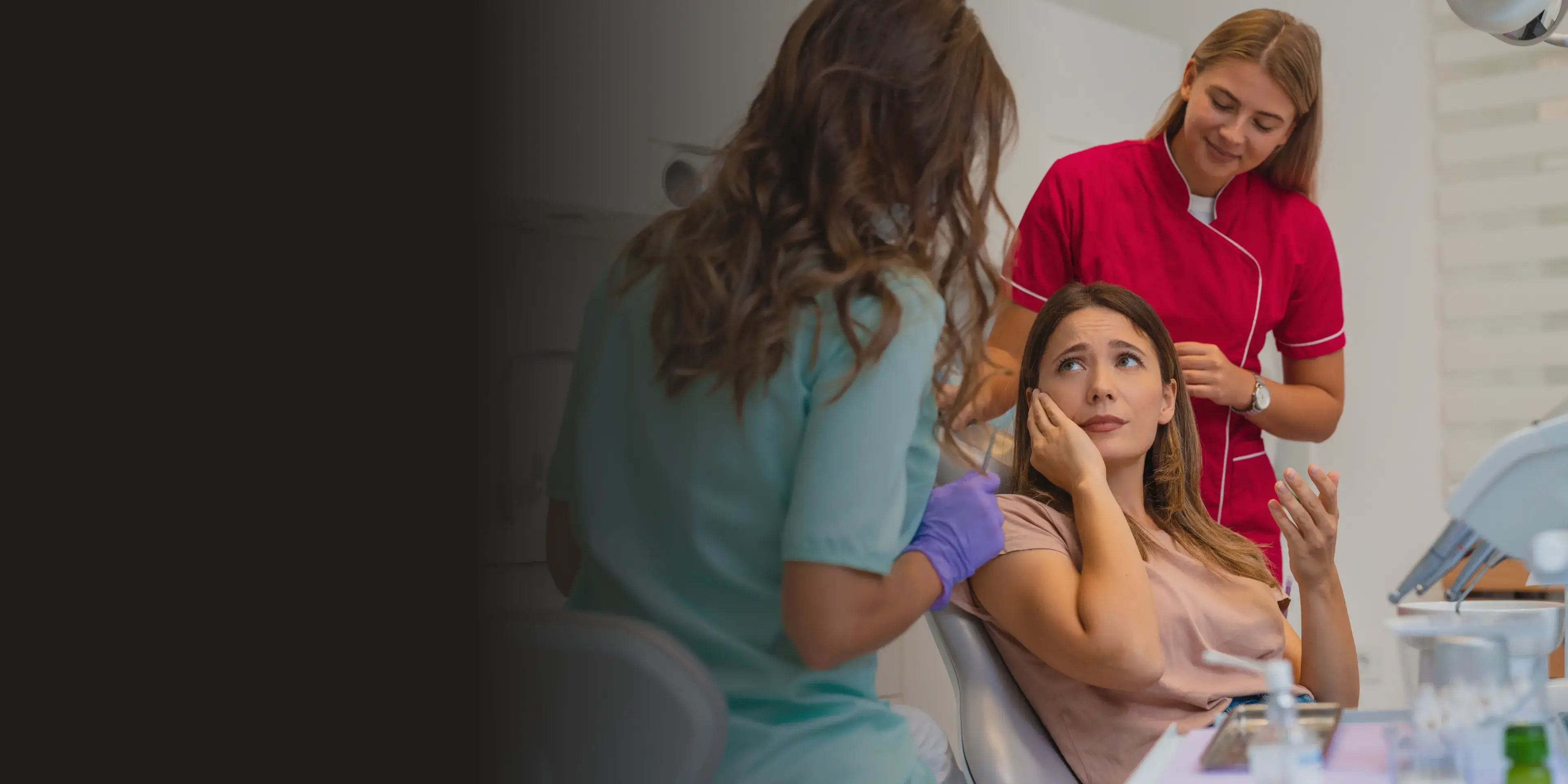 Woman seated in a dental chair holding her cheek in pain while a female dentist in teal scrubs prepares a syringe and a dental assistant in red scrubs stands nearby.