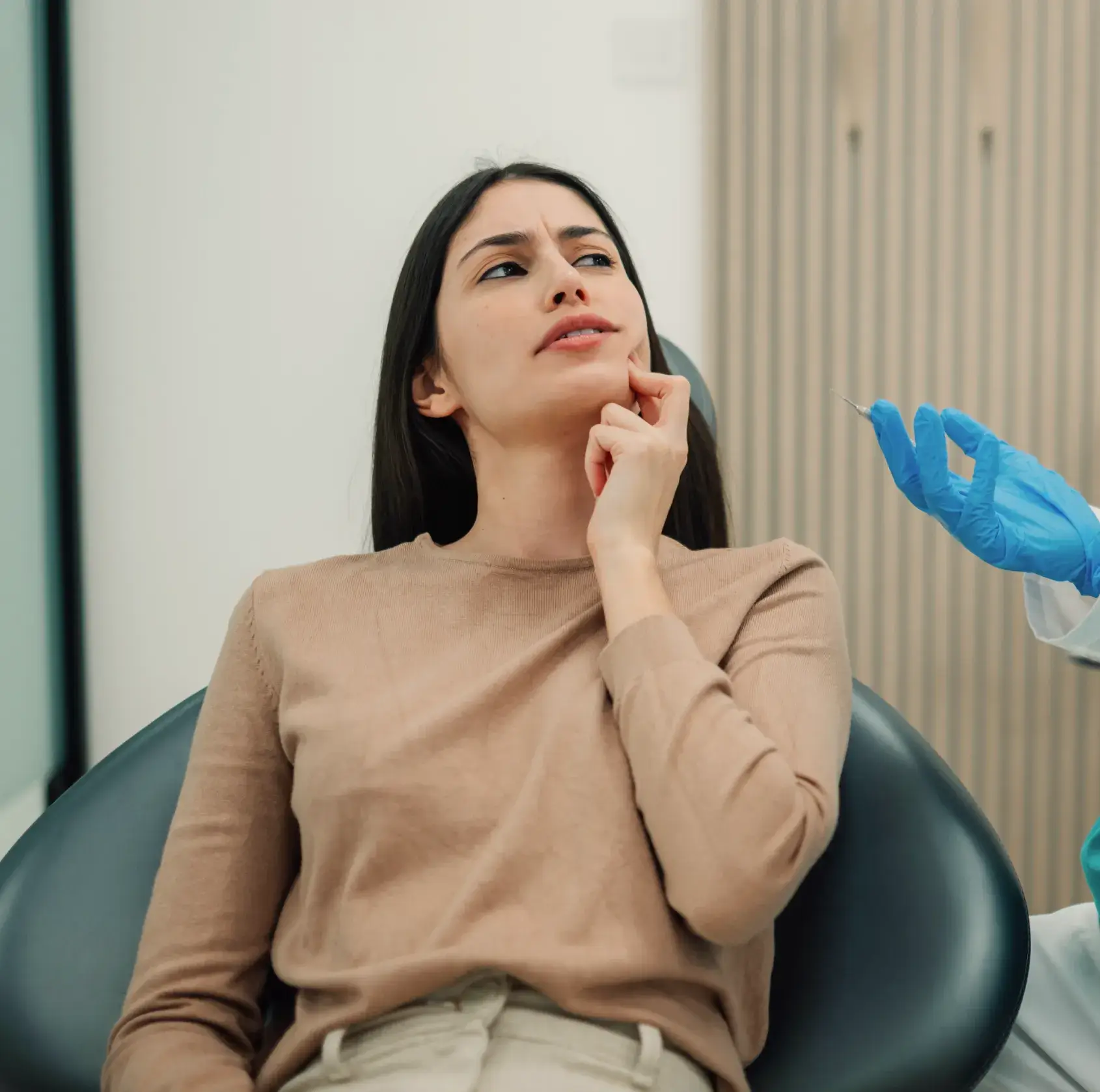 Woman sitting in a dental chair touching her cheek with a worried expression as a gloved hand holds a syringe nearby.