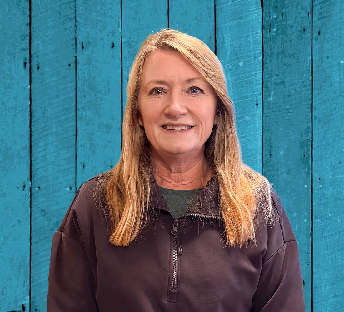 Our owner, Jeanette, wearing a dark zip-up jacket, standing against a turquoise wooden plank background.