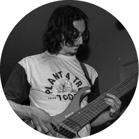 Man with medium-length hair and glasses playing an electric bass guitar.