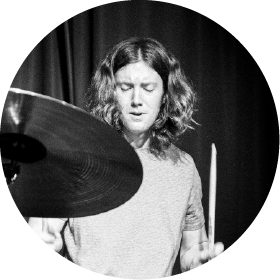 Person with shoulder-length hair playing drums, eyes closed, captured in black and white.