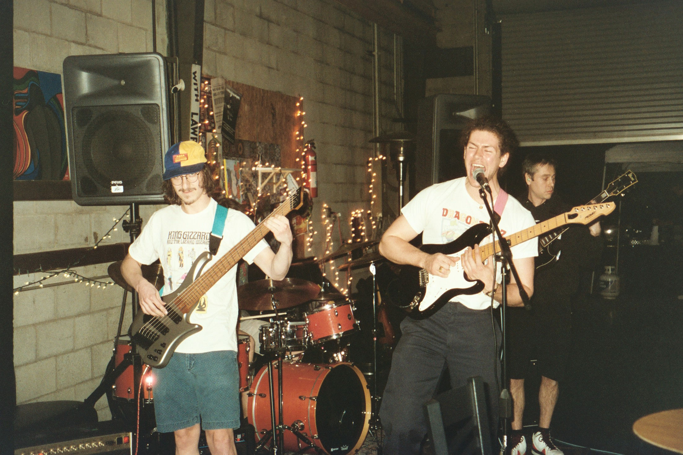 Three musicians performing indoors: a man with a bass guitar wearing a multicolored cap and King Gizzard T-shirt, a man singing and playing electric guitar, and another man in the background with an electric guitar.