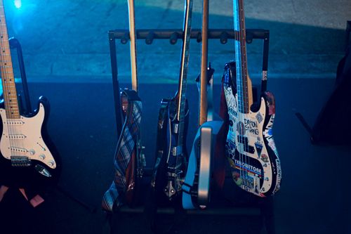 Close-up of several electric guitars and bass guitars on a rack, one bass guitar decorated with colorful stickers including text 'DAYTON STRONG'.