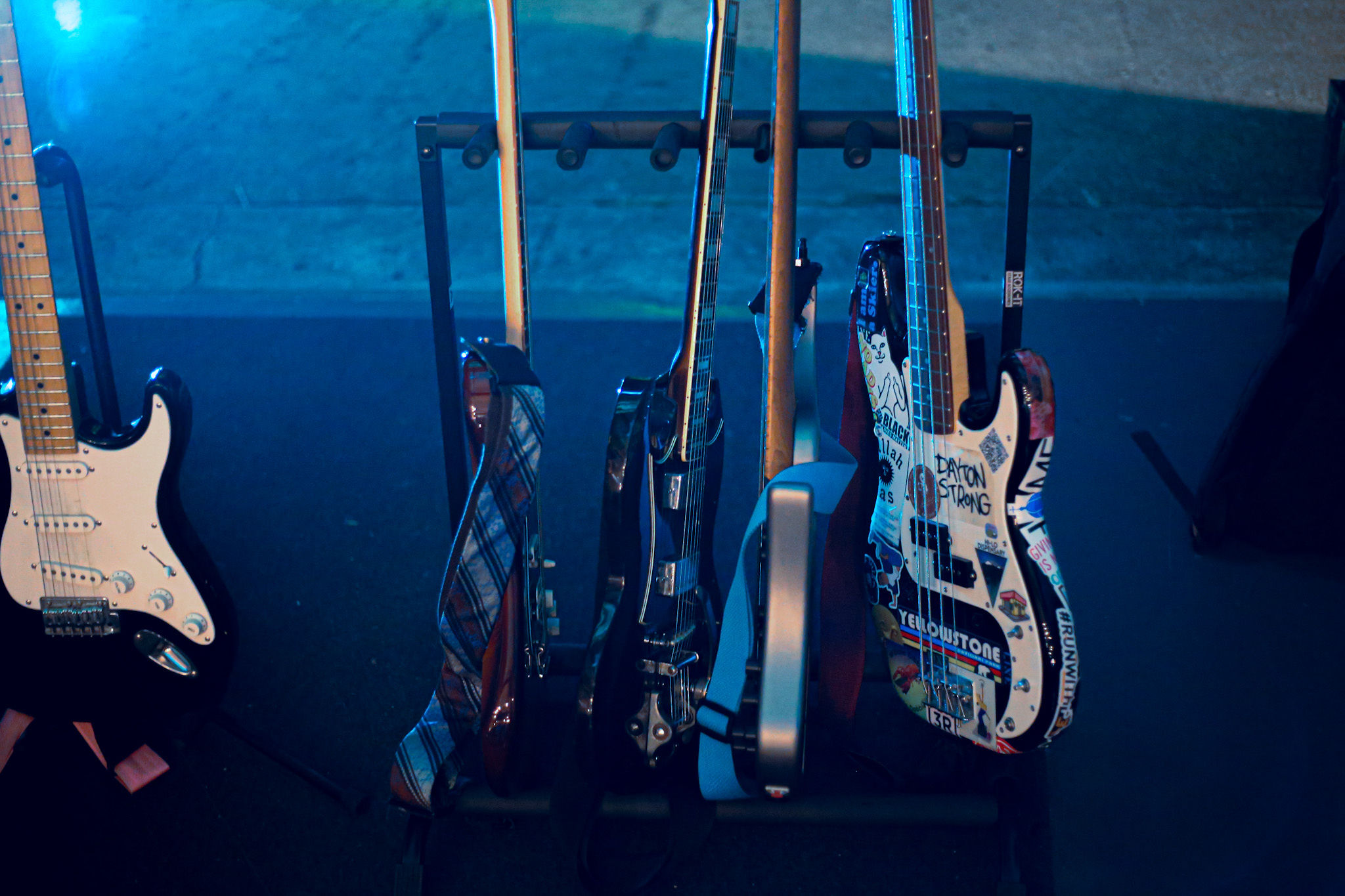 Close-up of several electric guitars and bass guitars on a rack, one bass guitar decorated with colorful stickers including text 'DAYTON STRONG'.