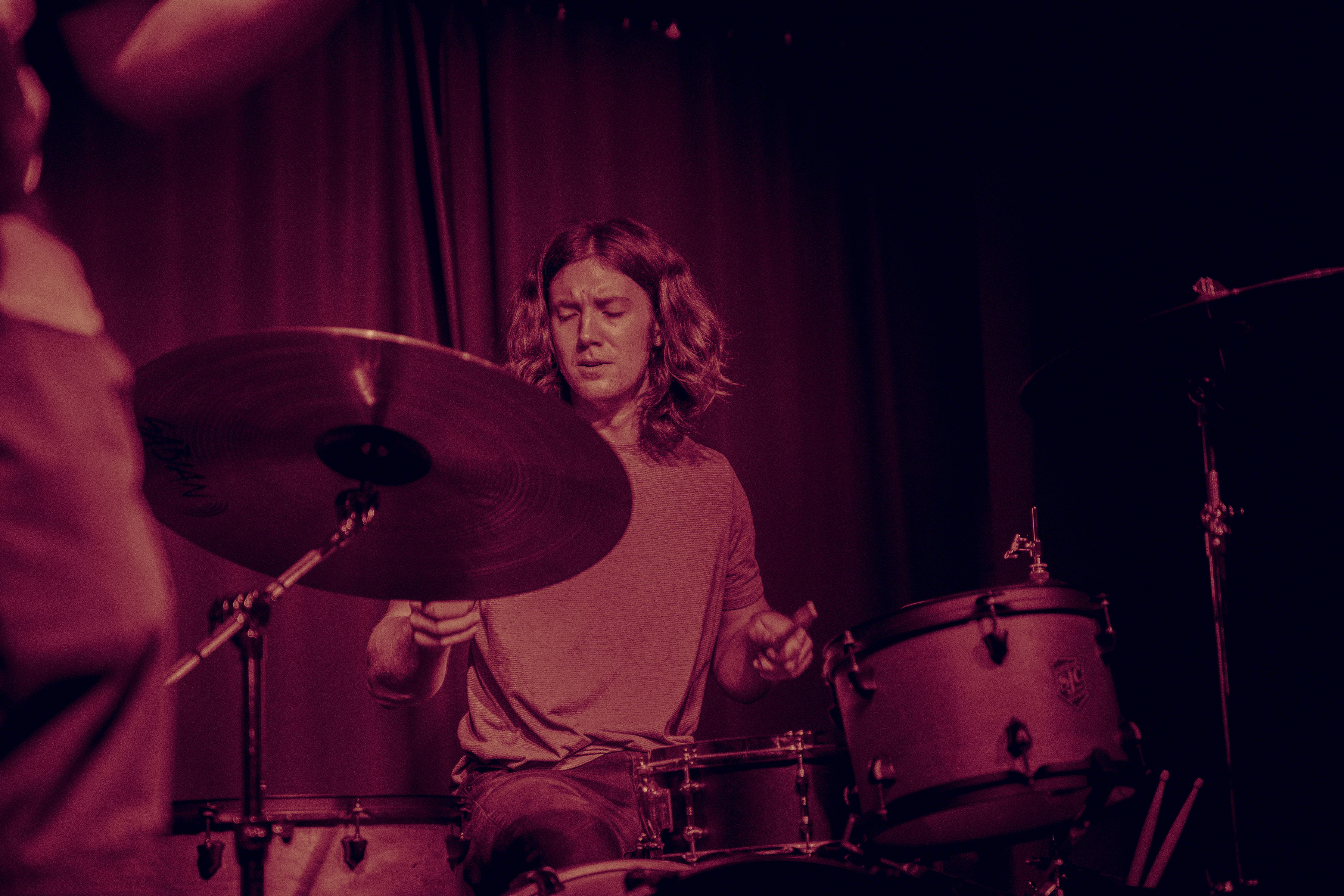 Young man with long hair playing a drum set on stage with focused expression.