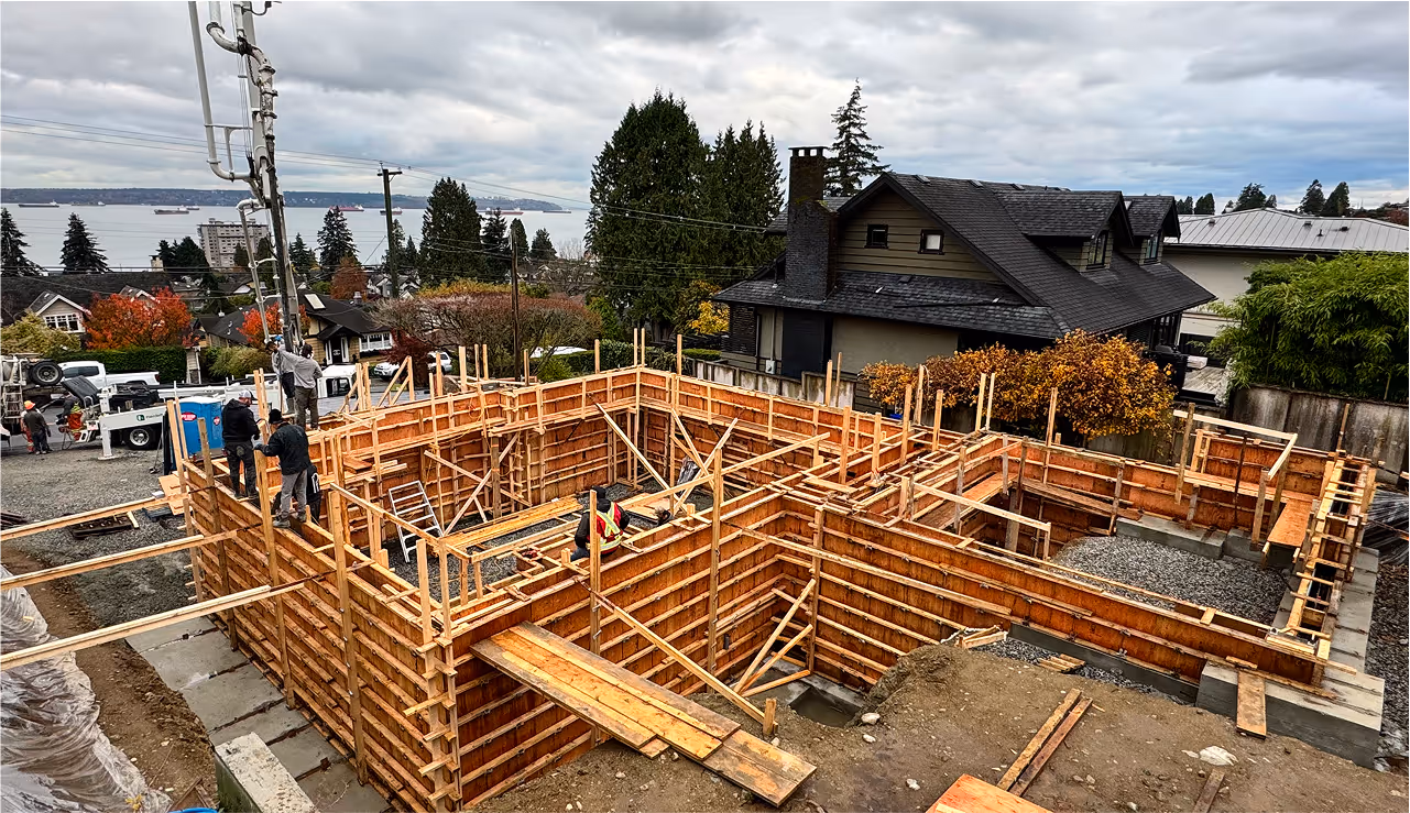 Construction site with wooden framing for a house foundation and workers on site, with a residential neighborhood and water body in the background under a cloudy sky.