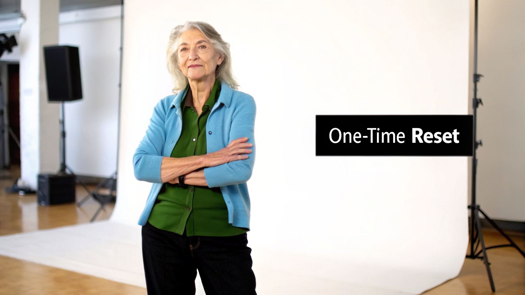 An older woman stands with arms crossed in a studio, next to a 'One-Time Reset' sign.