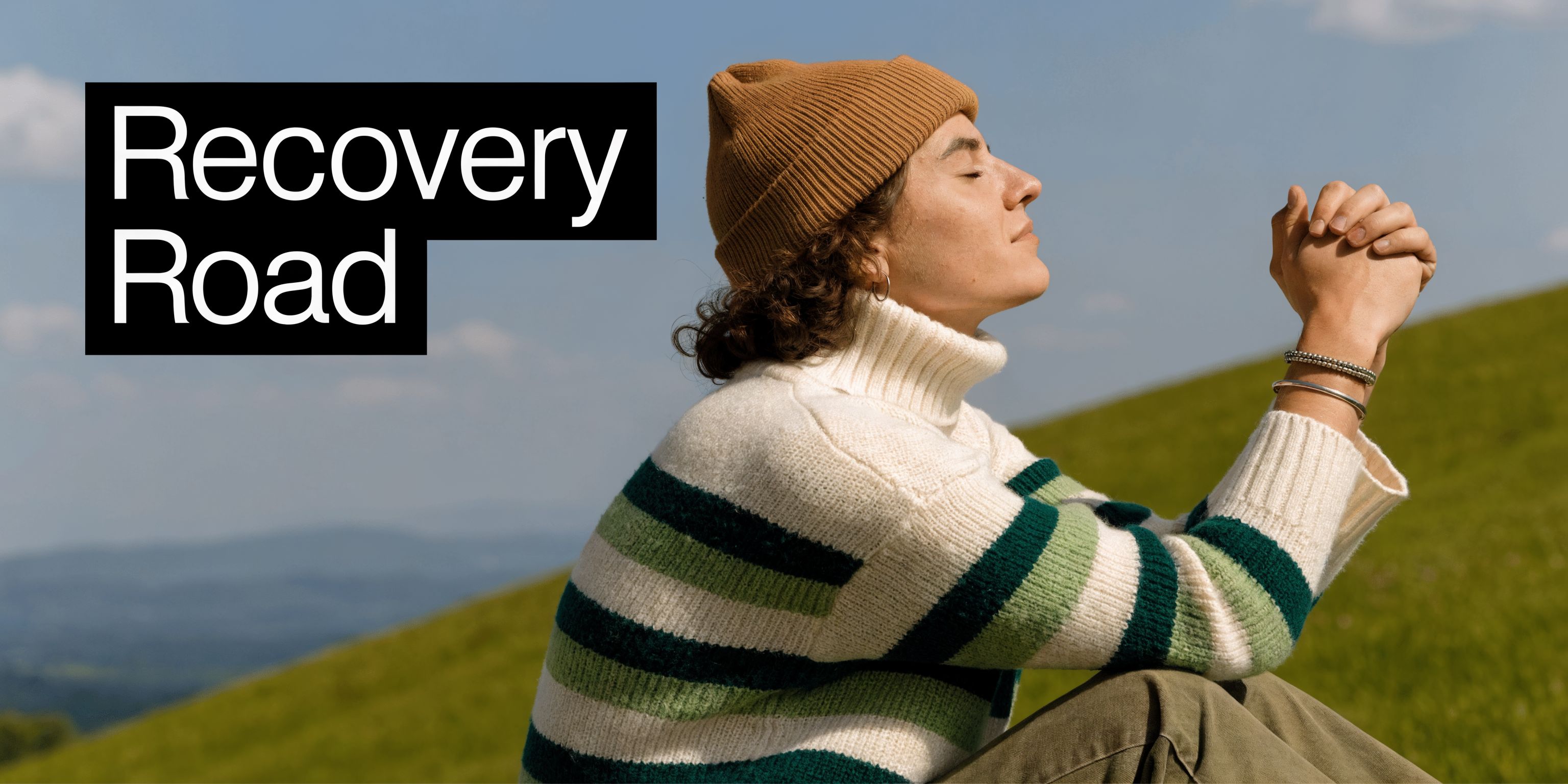 A person in a beanie and sweater sitting on a grassy hill, peacefully practicing mindfulness in nature.