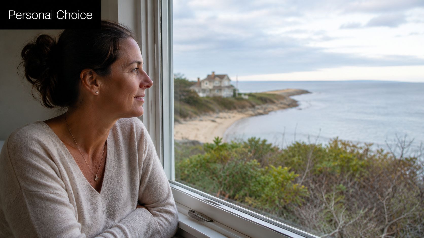A woman looks out a window at a scenic coastal view of a house by the ocean.