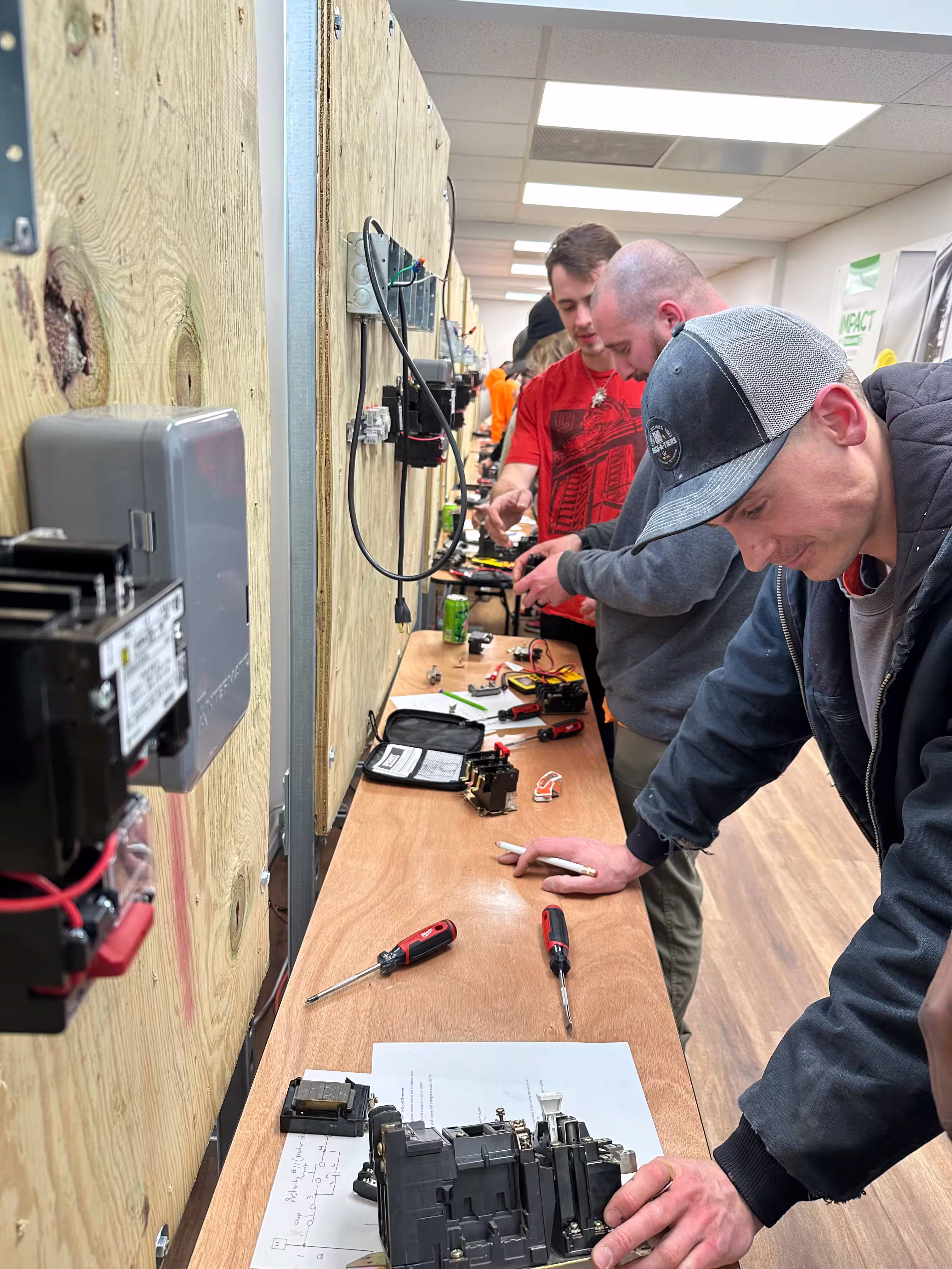 A group of Independent Electrical Contractors at a work bench covered with miscellaneous tools and parts, hard at work.