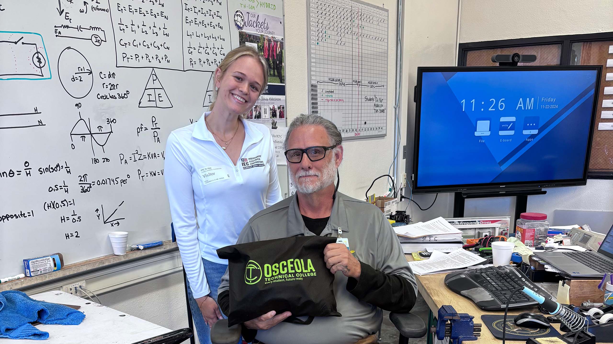 A young woman from the IEC poses for a photo with a teacher in front of a whiteboard covered with formulas and equations.