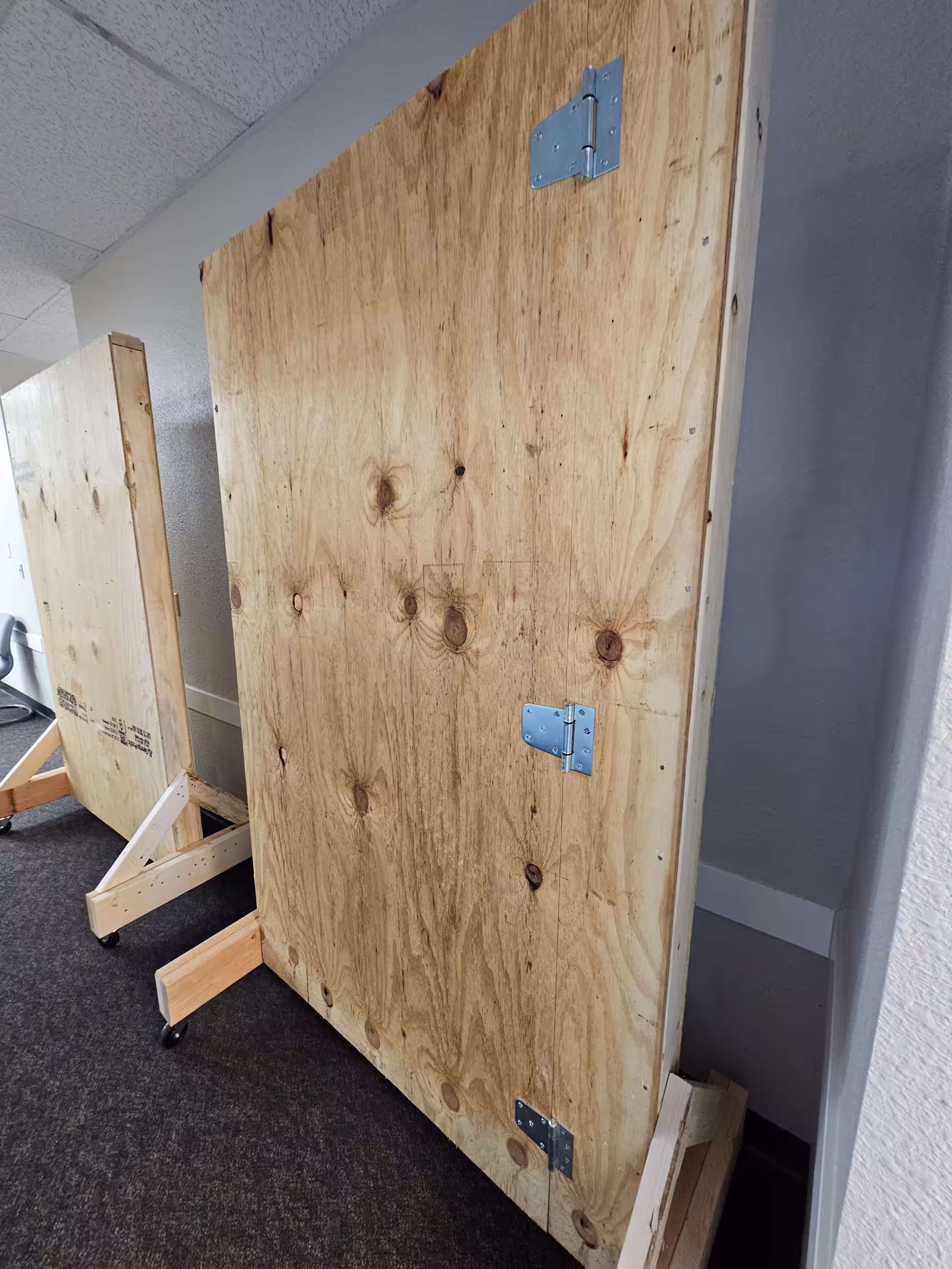 An image of a plywood board standing on its own with pencil marks and door hinges drilled into it.