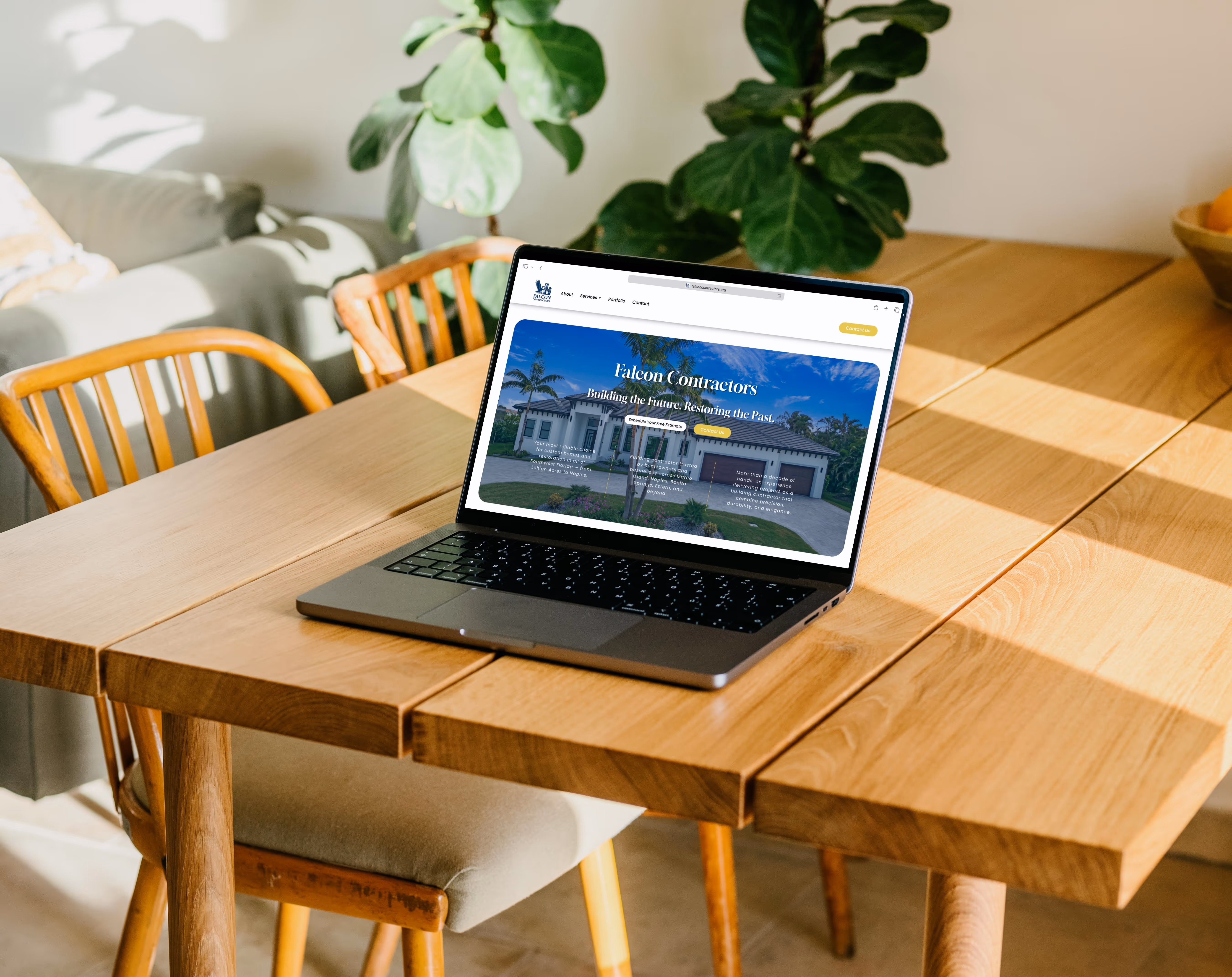 Laptop displaying Falcon Contractors website with house image, placed on wooden dining table with chairs and plants in background.