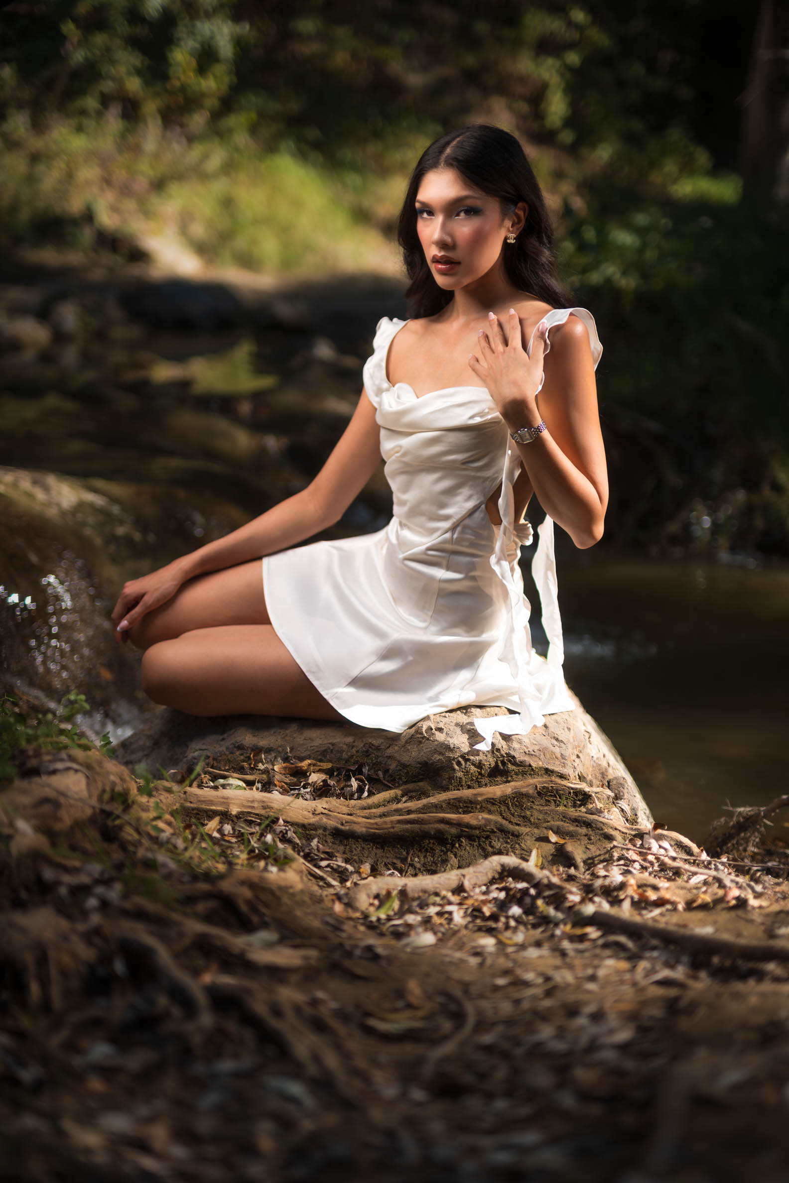Athena Poulos seated on a rock beside a creek in a white satin dress during an outdoor fashion editorial in Malibu, California.