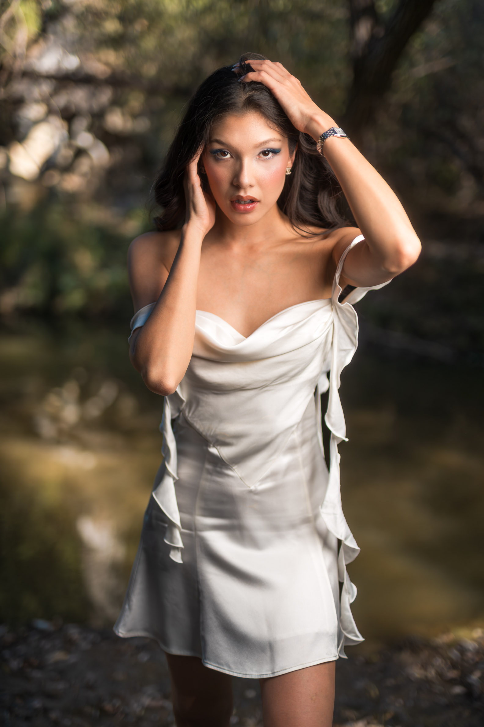 Close-up editorial portrait of Athena Poulos in a white satin dress, looking toward the camera with her hands framing her face.